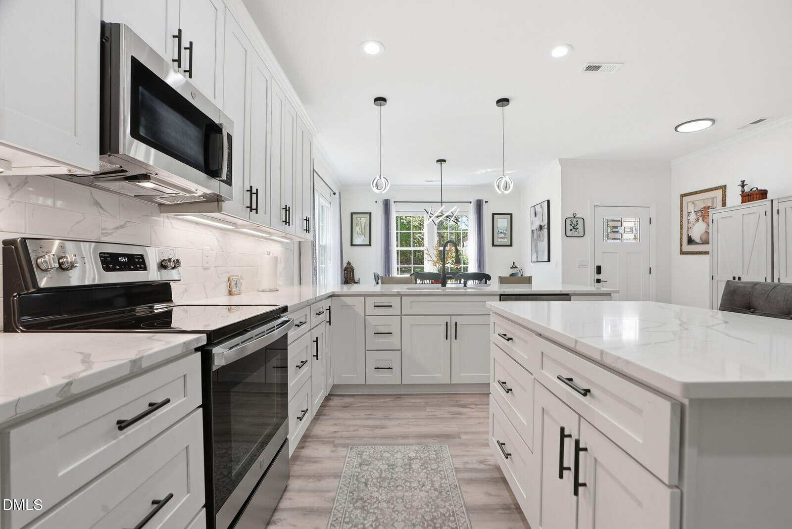 Bright modern kitchen with white cabinets, marble countertops, stainless steel appliances, pendant lights, and a large island. A dining area with windows and wall art is visible in the background.