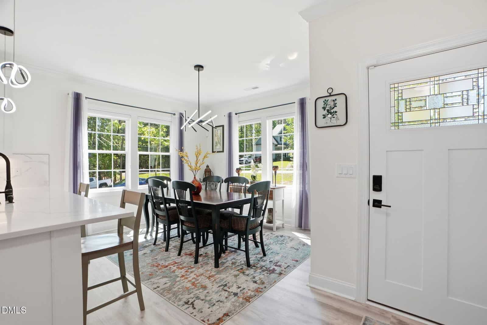 Bright dining area with large windows, a rectangular table with eight chairs, and a modern chandelier. A colorful rug is under the table; the entry door with glass detail is on the right. Light and airy atmosphere.