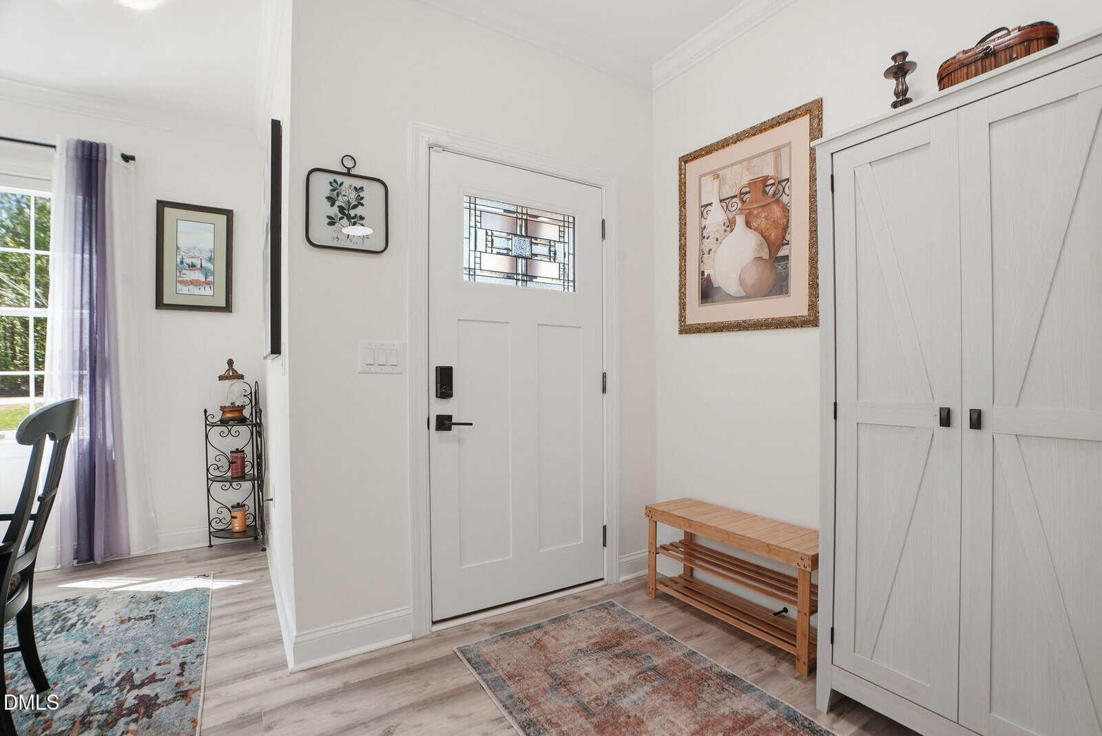 Bright entryway with a white door, stained glass window, wooden bench, large wardrobe, wall art, and a small rug on light wood floors. A sunlit window with purple curtains and decor are visible in the adjacent room.