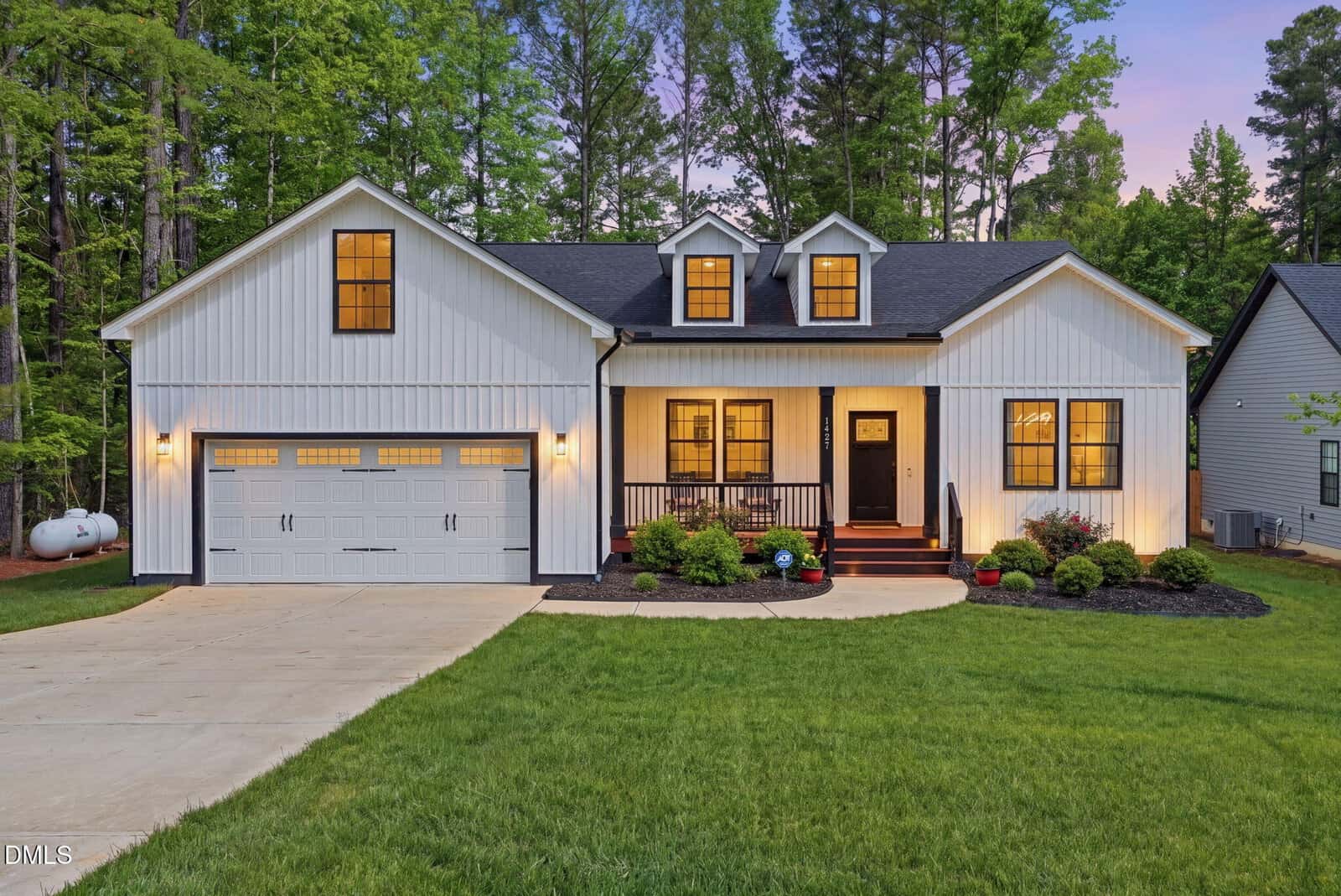 A modern white house with black trim at 1427 Sagamore Drive, Louisburg features a double garage, front porch with chairs, landscaped yard, and warm lights glowing at dusk. Tall trees and neighboring houses complete the inviting scene.