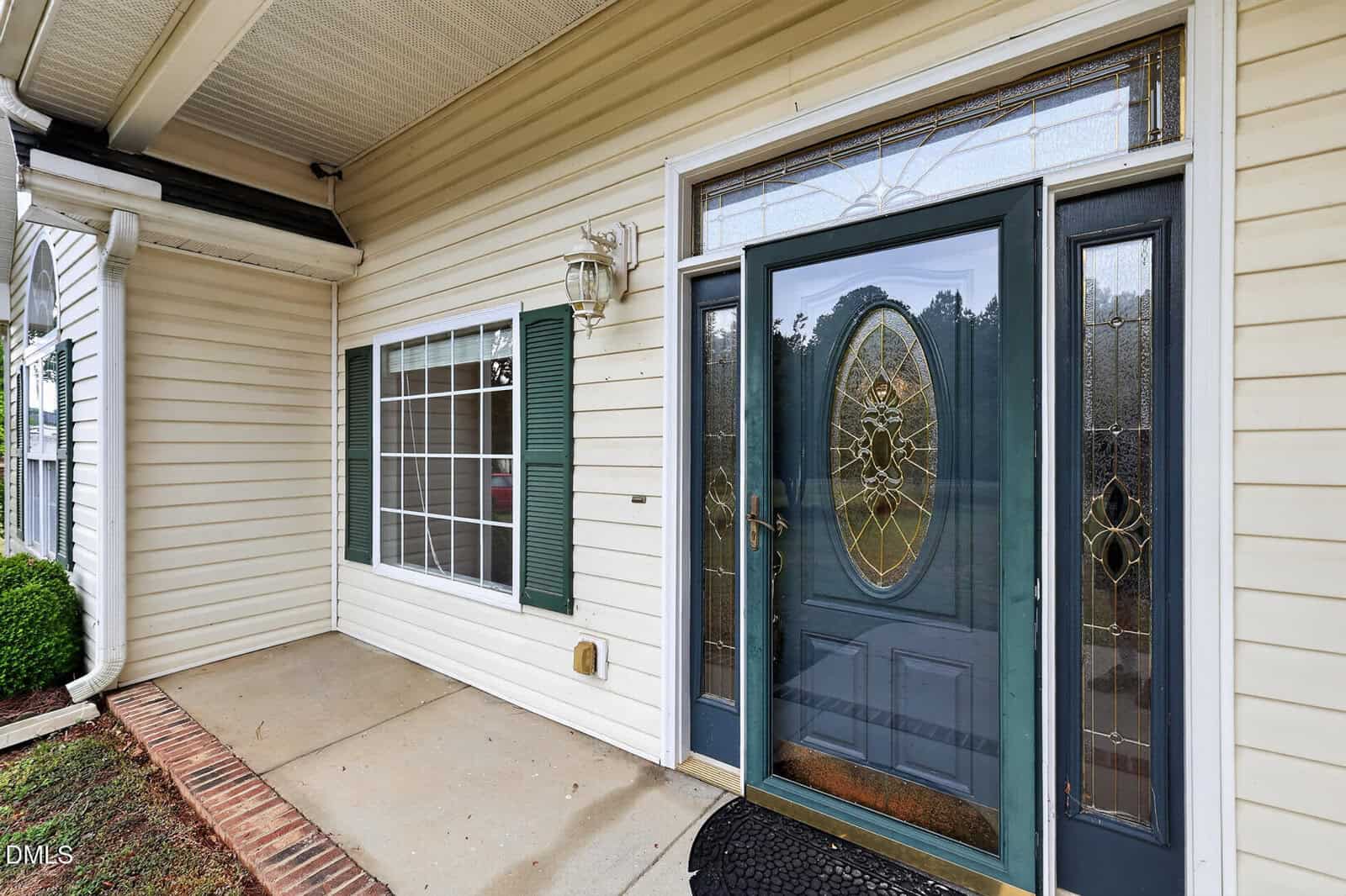 Front entryway of a house with beige siding, a large window with green shutters, a decorative glass front door, a small porch area, and a wall-mounted light fixture.