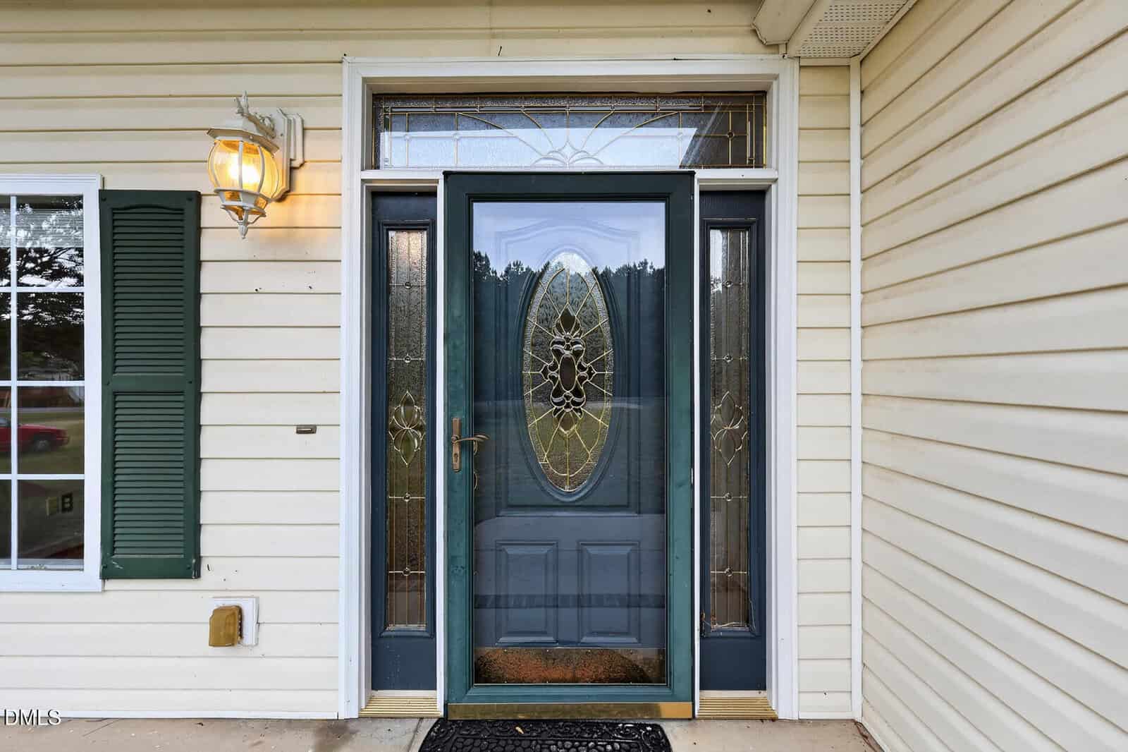 A front door with an oval-shaped, decorative glass panel, flanked by two matching glass sidelights, cream-colored siding, a window with green shutters, and a lantern-style porch light.