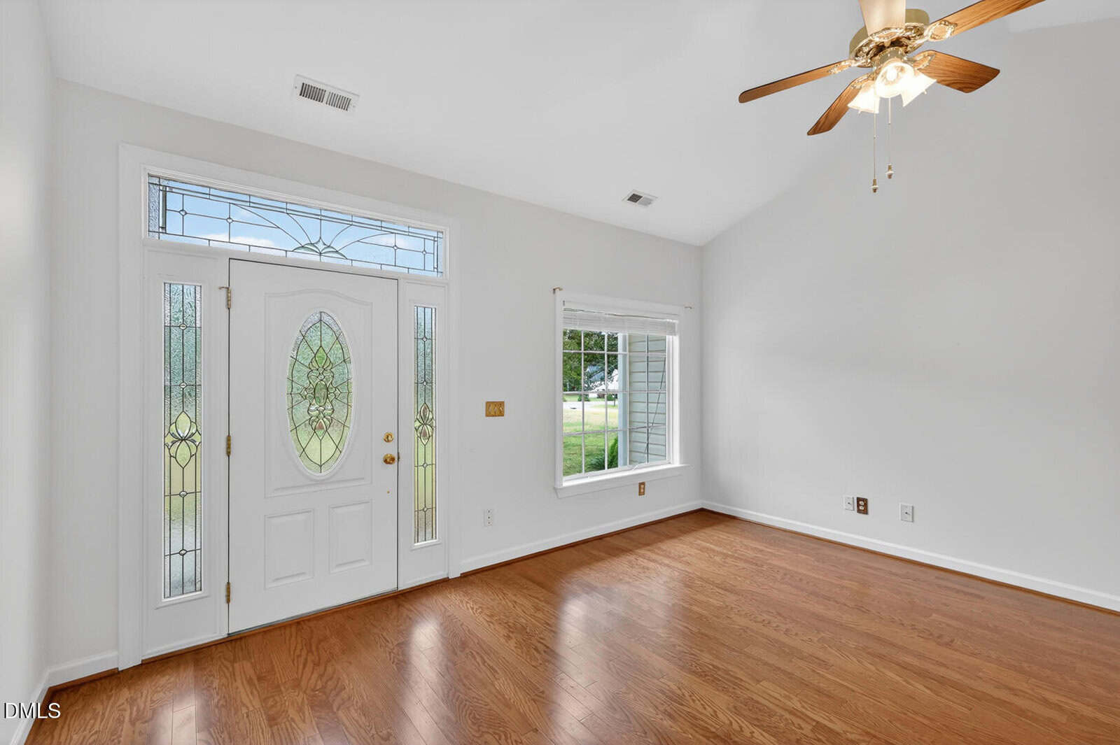 Bright, empty room with hardwood floors, white walls, a ceiling fan with lights, a large window, and a decorative white front door with glass panels. Natural light fills the space.