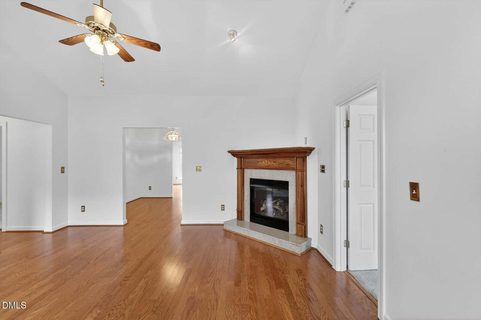 A living room with wood flooring, a ceiling fan, and a corner fireplace with a wooden mantel. There are open doorways leading to other rooms, and white walls throughout the space.
