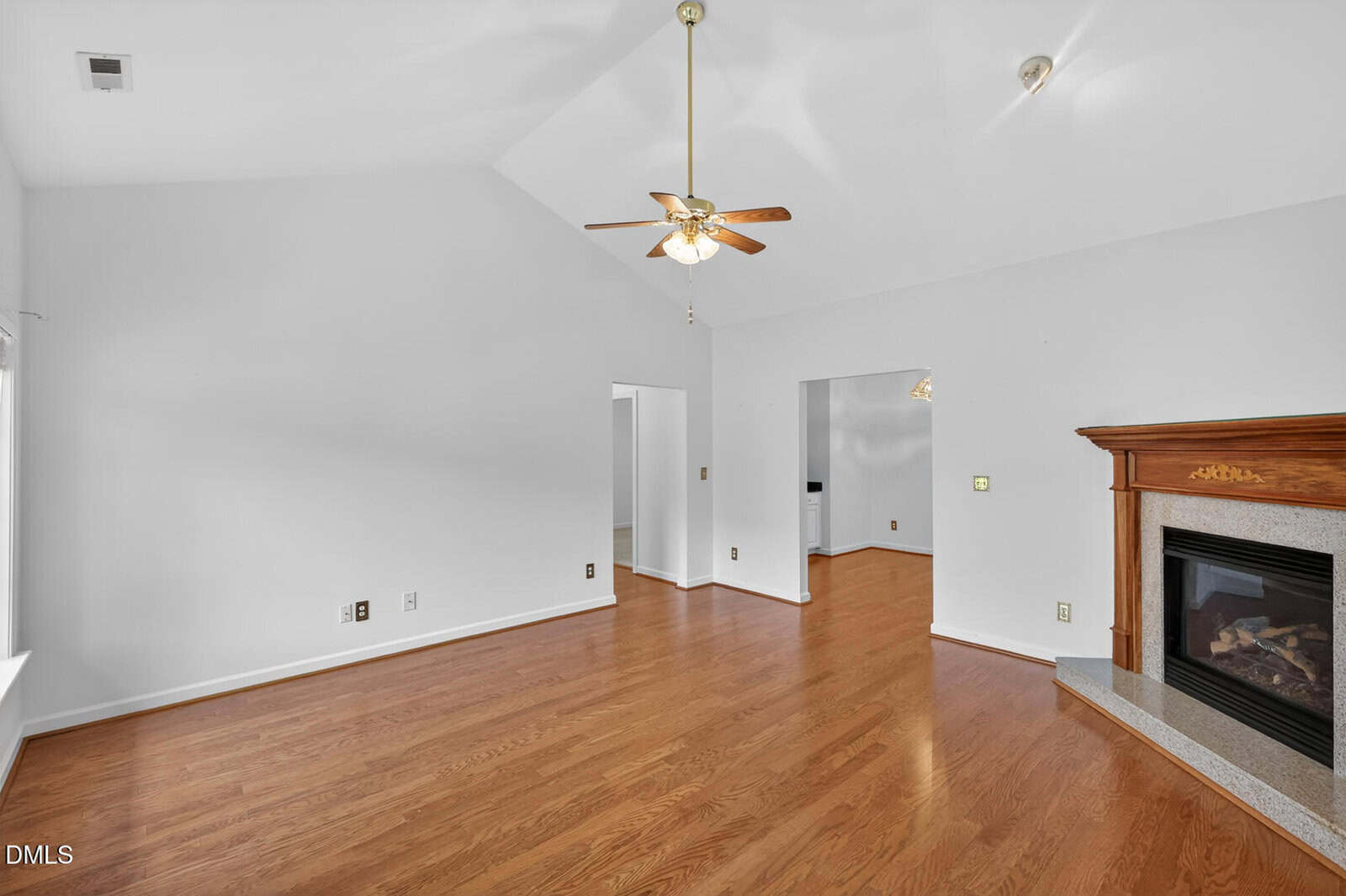 Bright living room with vaulted ceiling, wooden floors, white walls, a ceiling fan with light, and a corner fireplace with a wooden mantle. Open doorways lead to adjacent rooms.