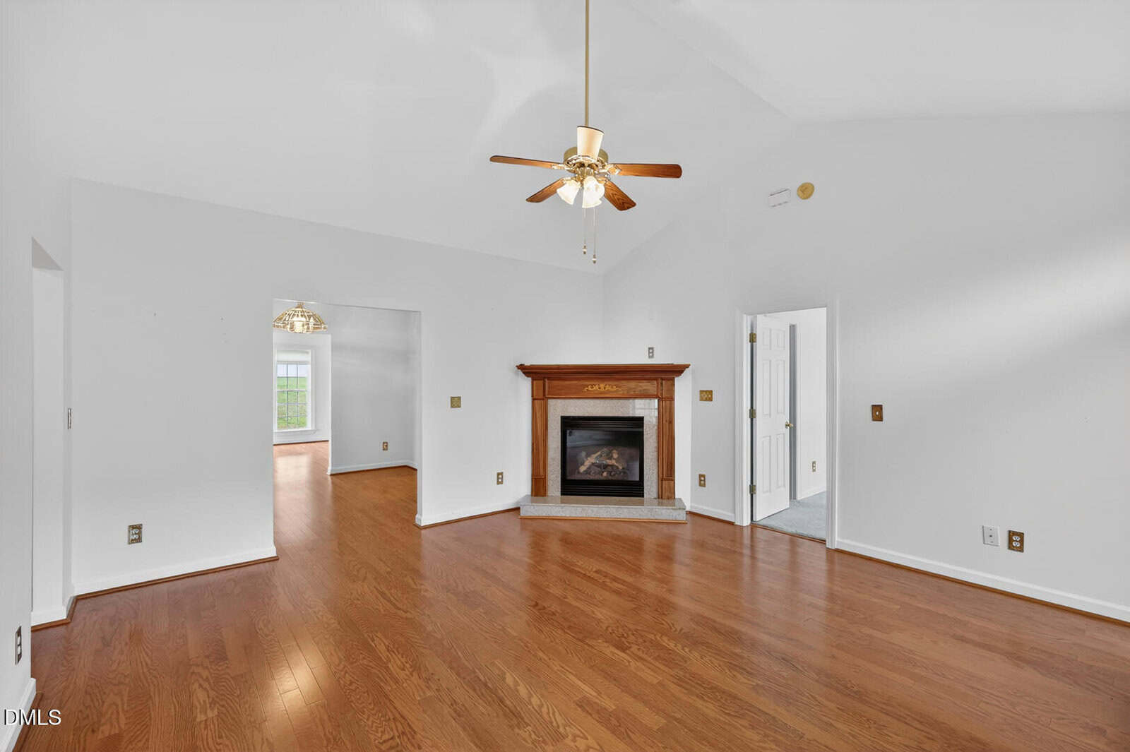 A spacious living room with hardwood floors, a wooden fireplace, white walls, a ceiling fan with lights, and open doorways leading to adjoining rooms.