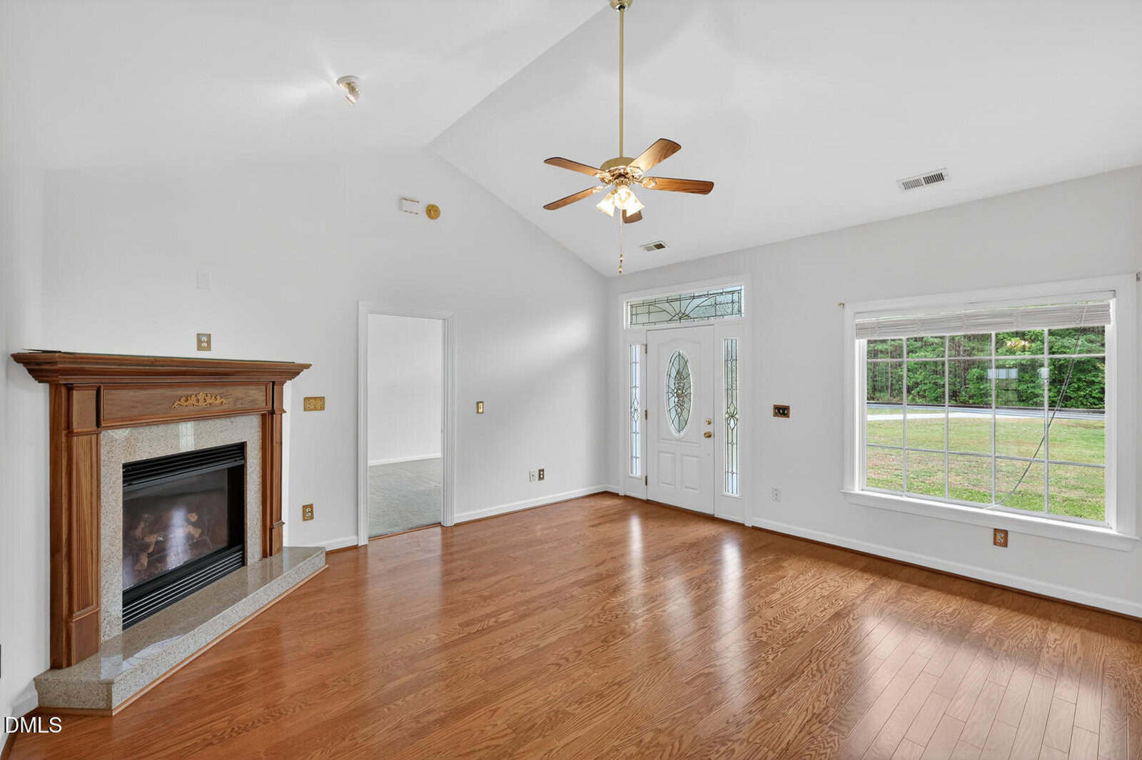 Bright living room with hardwood floors, white walls, a ceiling fan, decorative fireplace, large window, and front door with glass panels. An open doorway leads to another room.