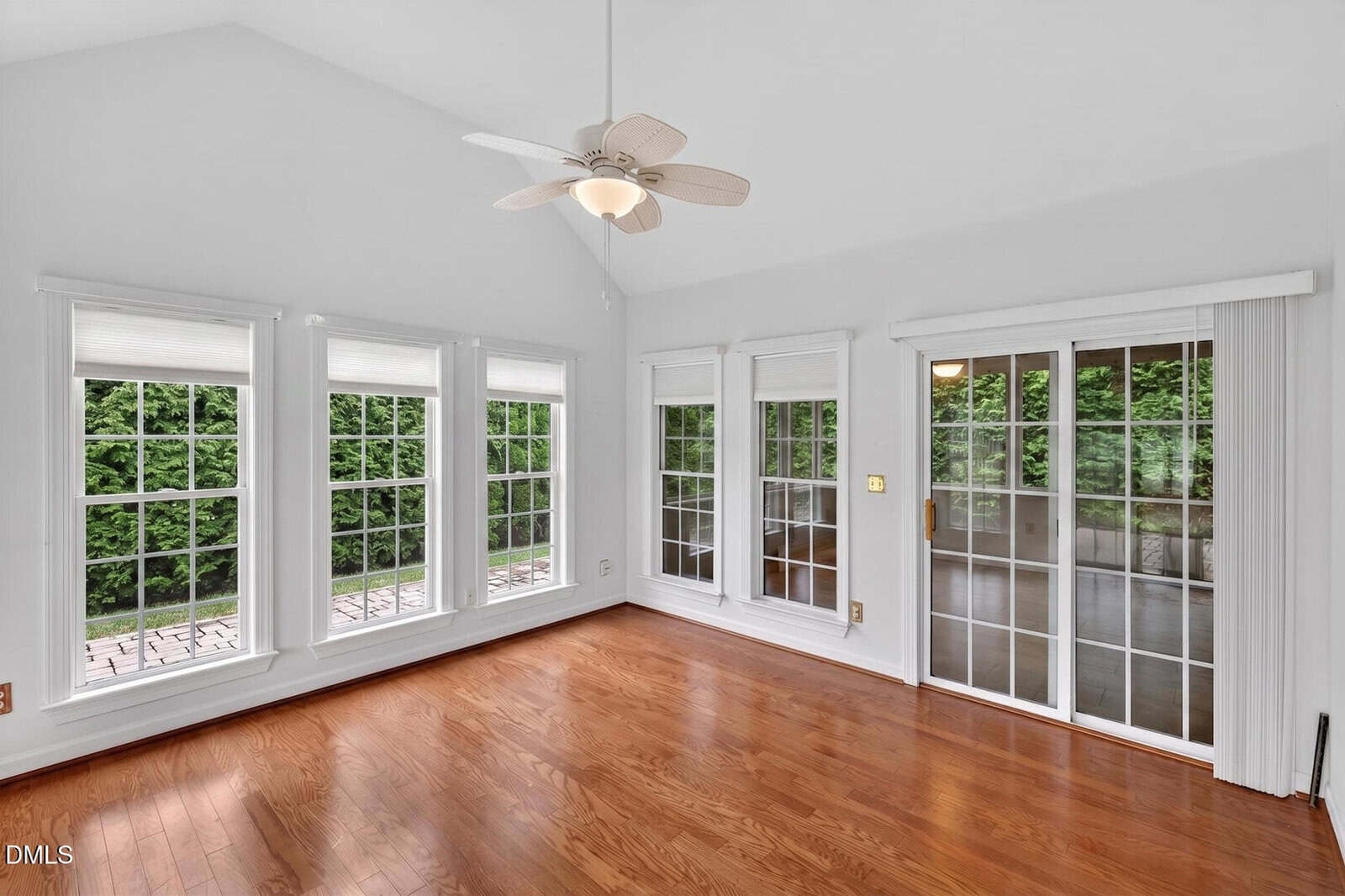 A bright room with large windows, a glass sliding door, hardwood floors, white walls, and a ceiling fan. Greenery is visible outside through the windows and door.