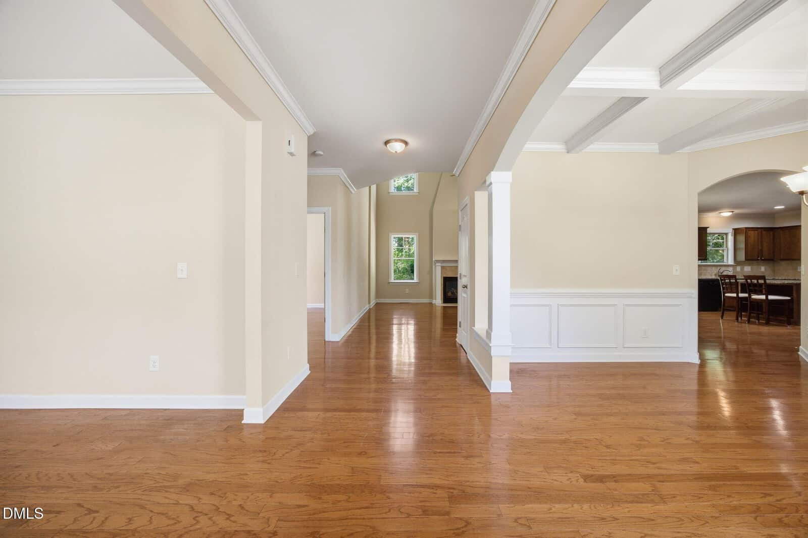 Bright, open hallway with light beige walls, white trim, and hardwood floors. Archways separate dining and living areas, and windows provide natural light. Kitchen is visible in the background.