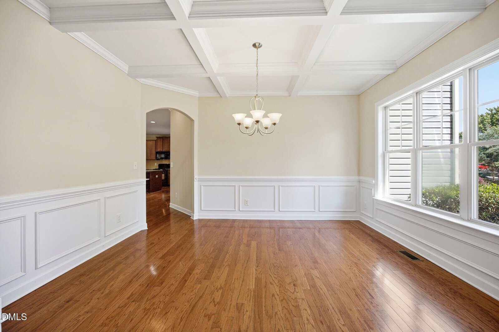 Bright dining room with hardwood floors, large window, white wainscoting, beige walls, coffered ceiling, and a modern chandelier. An open doorway leads to another room, and greenery is visible outside.