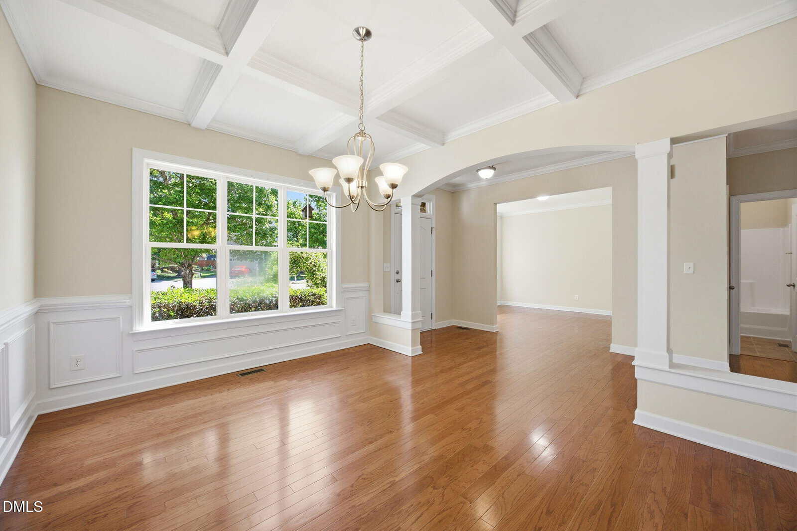 Bright dining area with wood floors, coffered ceiling, large window, and chandelier opens into a spacious living room with white trim and neutral walls. Door to the right leads to another room.