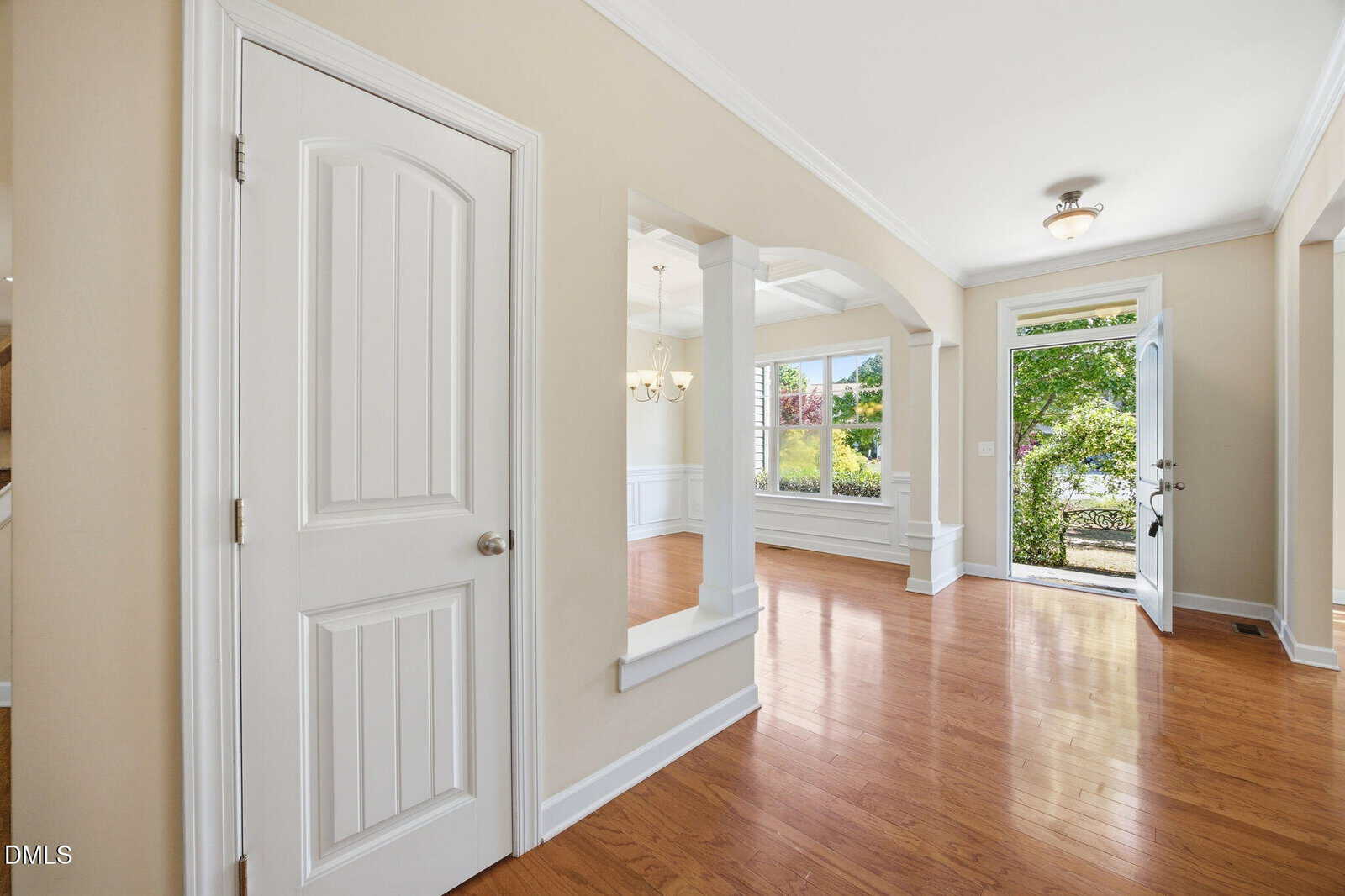 Bright entryway with light hardwood floors, beige walls, white trim, a closed paneled door, open archways, and a glass front door showing greenery outside. Large windows bring in natural sunlight.