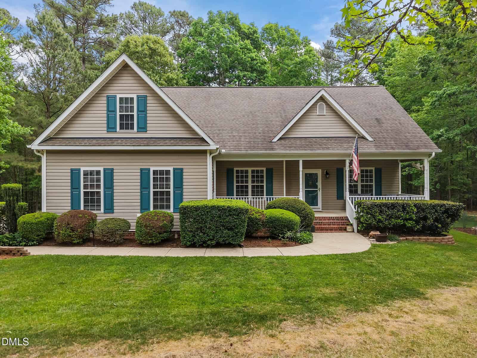 A beige two-story house with green shutters, a covered front porch, and an American flag. Well-maintained shrubs line the walkway, and tall trees are visible in the background.