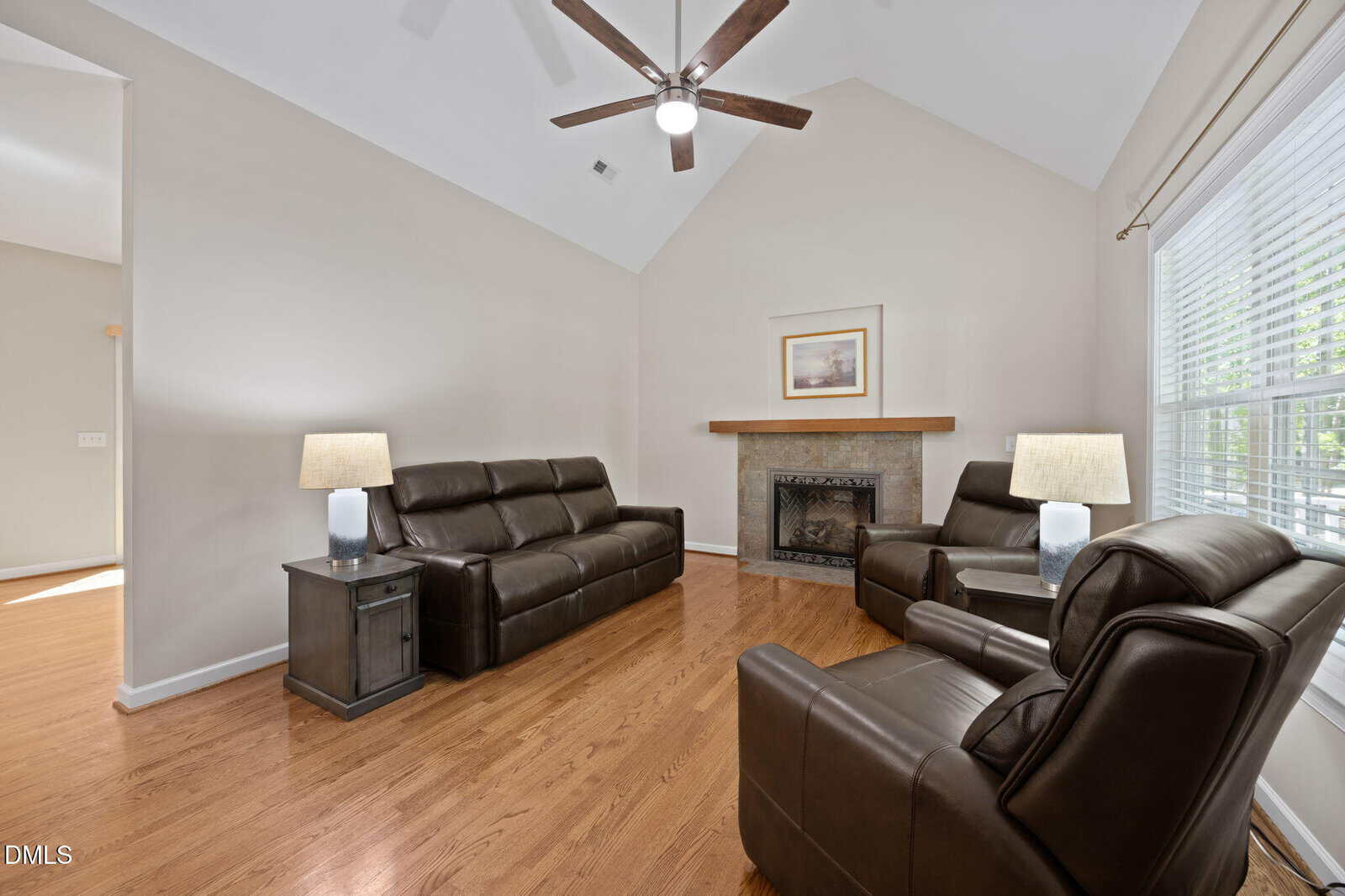 A living room with two brown leather sofas and an armchair, a fireplace with a framed picture above it, two side tables with lamps, hardwood floors, and large windows with blinds letting in natural light.