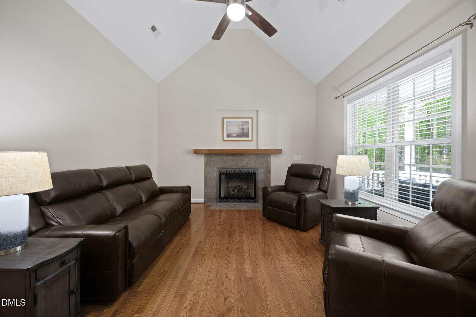 A living room with vaulted ceiling, fireplace, two brown leather sofas, a matching armchair, wooden floor, two side tables with lamps, a large window with blinds, and a framed picture above the mantel.