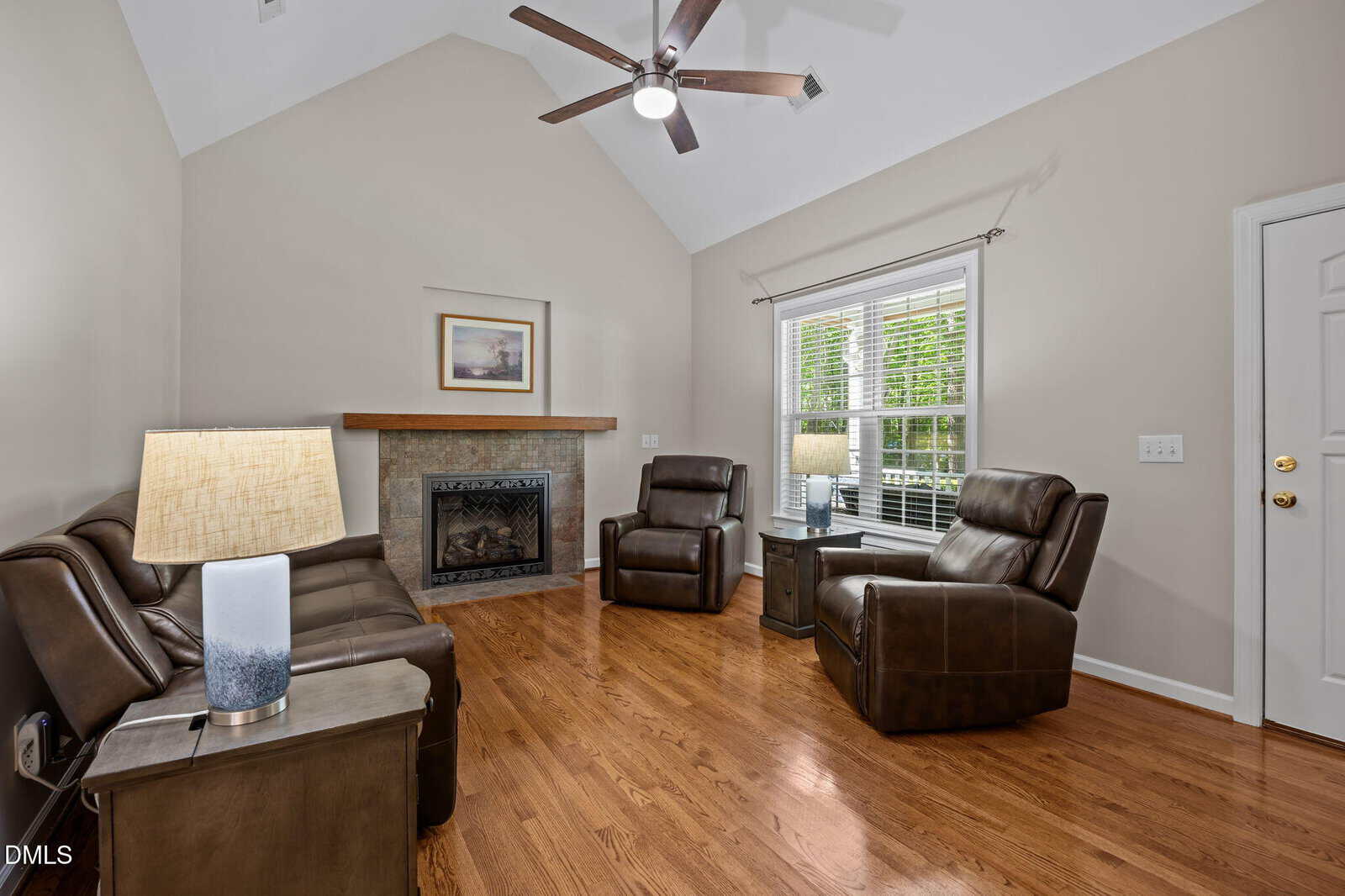 Living room with hardwood floors, brown leather seating, a fireplace with a mantle, side tables with lamps, a ceiling fan, and a large window letting in natural light.