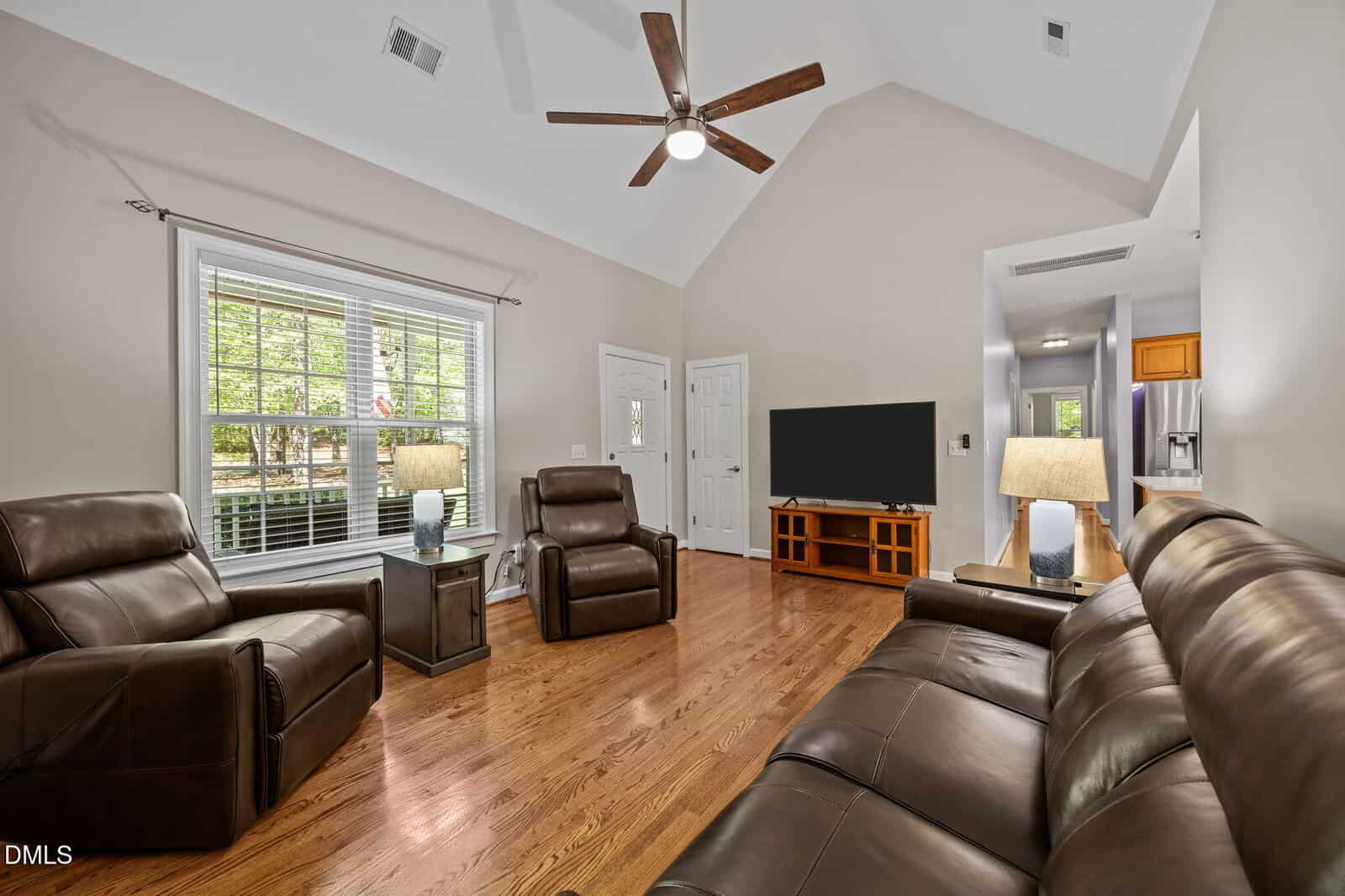 Living room with brown leather sofa and recliners, wood floors, a large window, TV on a stand, ceiling fan, lamps, and view into kitchen with stainless appliances. Walls are light-colored and ceiling is vaulted.