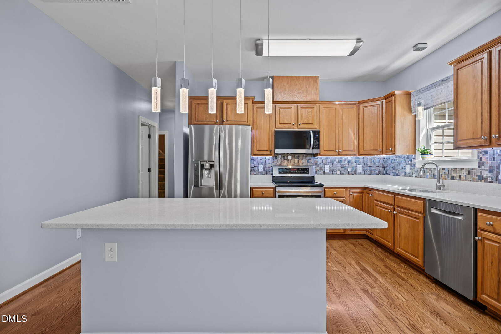 Modern kitchen with wood cabinets, stainless steel appliances, quartz island, tile backsplash, pendant lights, light blue walls, and hardwood floors. A window is above the sink, and a doorway is visible in the background.