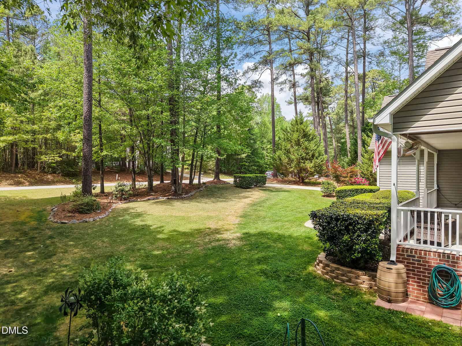 A well-kept backyard with green grass, trees, manicured bushes, and a small brick patio. An American flag hangs from the house, and a quiet, wooded street is visible in the background.