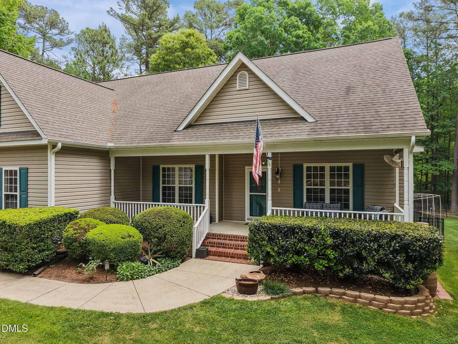 A beige single-story house with a covered front porch, white railings, an American flag, green shutters, red brick steps, neatly trimmed bushes, and a well-maintained lawn surrounded by trees.