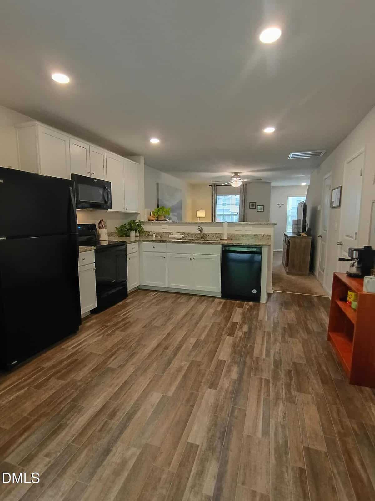 Modern kitchen with wood-style flooring, black appliances, white cabinets, a granite countertop, recessed lighting, and a small open shelving unit on the right. A living area is visible beyond the kitchen.