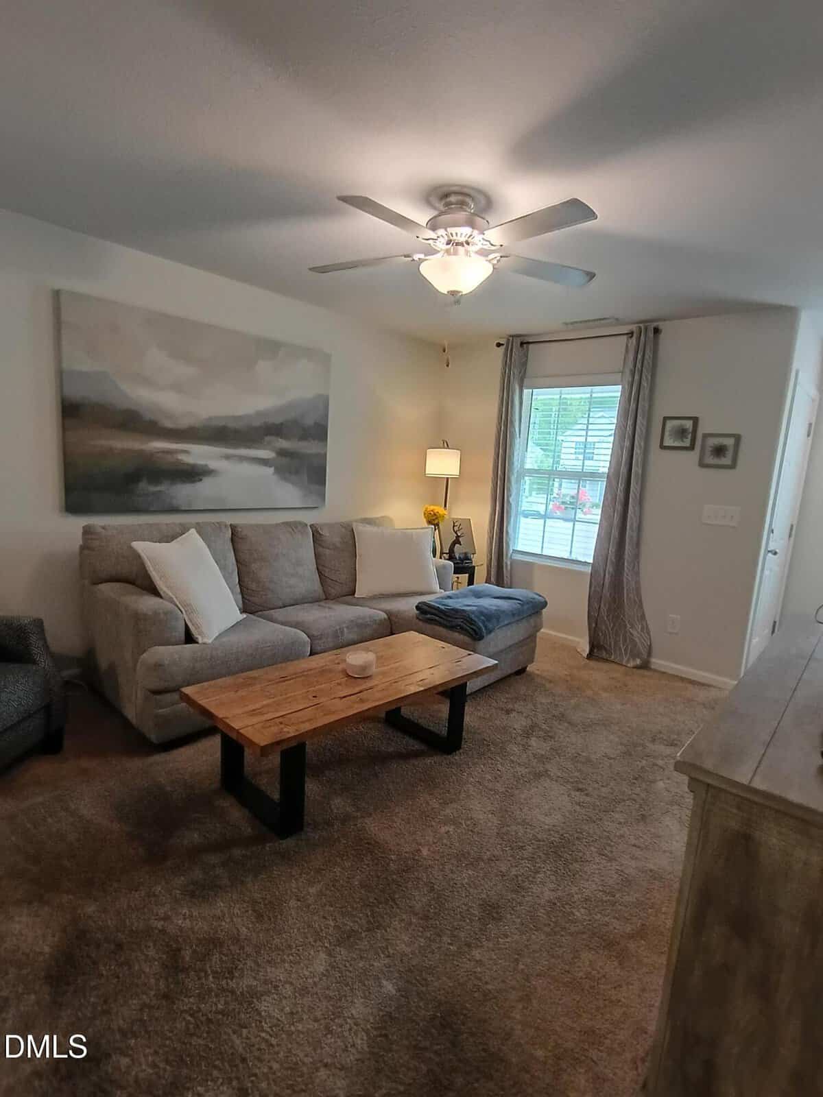 Cozy living room with a gray sofa, wooden coffee table, landscape painting, and window with sheer curtains. Ceiling fan and soft lighting create a welcoming atmosphere.