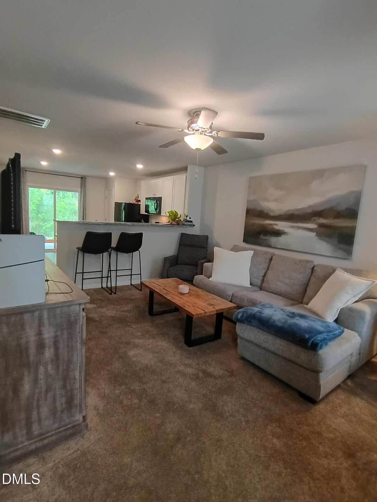 Living room with gray sectional sofa, wooden coffee table, two bar stools at a kitchen counter, large wall art, ceiling fan, TV on a stand, and sliding glass doors leading to a green outdoor area.