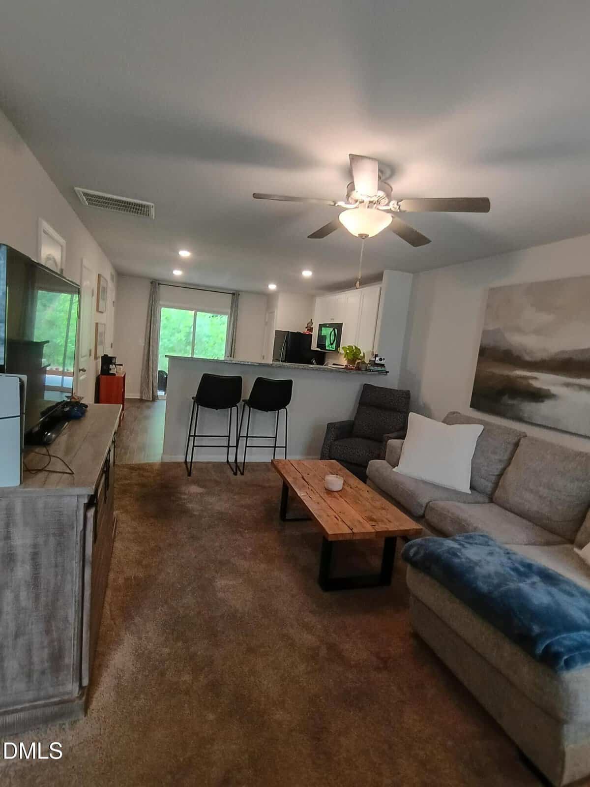 A cozy living room with brown carpet, a gray sectional sofa, a wooden coffee table, and a TV on a stand. In the background, there's a kitchen with two bar stools and a ceiling fan overhead.