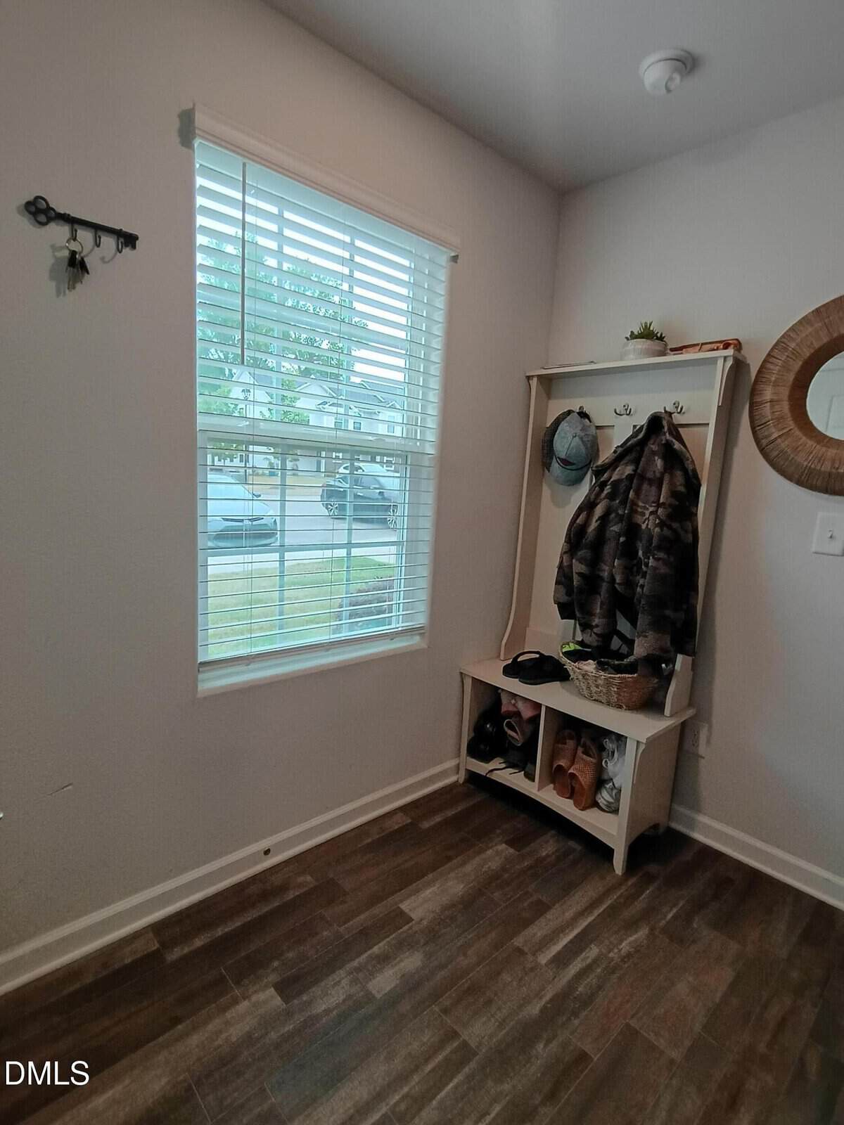 A small entryway with a window, wooden bench with hooks, coats, hats, a basket, and shoes. The floor is dark wood, and a round mirror hangs on the wall. Sunlight streams through the window blinds.