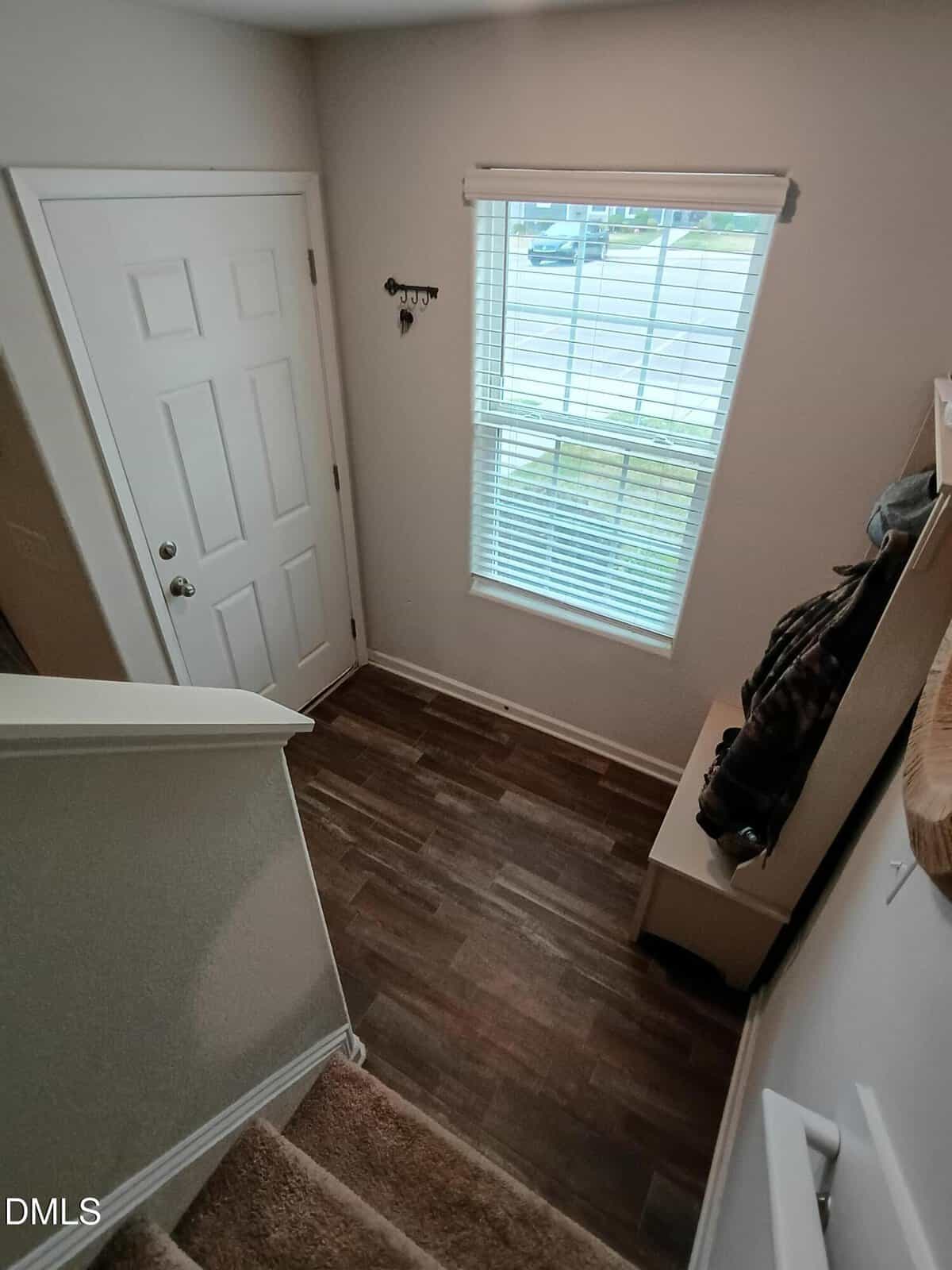A view from above of a small entryway with a white door, a window with blinds, a wooden floor, and a bench with jackets and bags. Stairs with carpet lead down to the entry area.