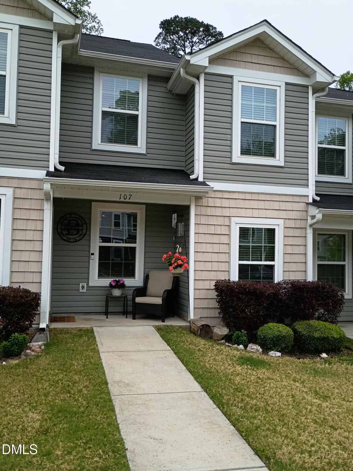 A two-story townhouse with gray siding, white trim, and tan shingles. The front porch has a black chair, potted flowers, and a hanging flower basket. A concrete path leads across a green lawn to the front door.