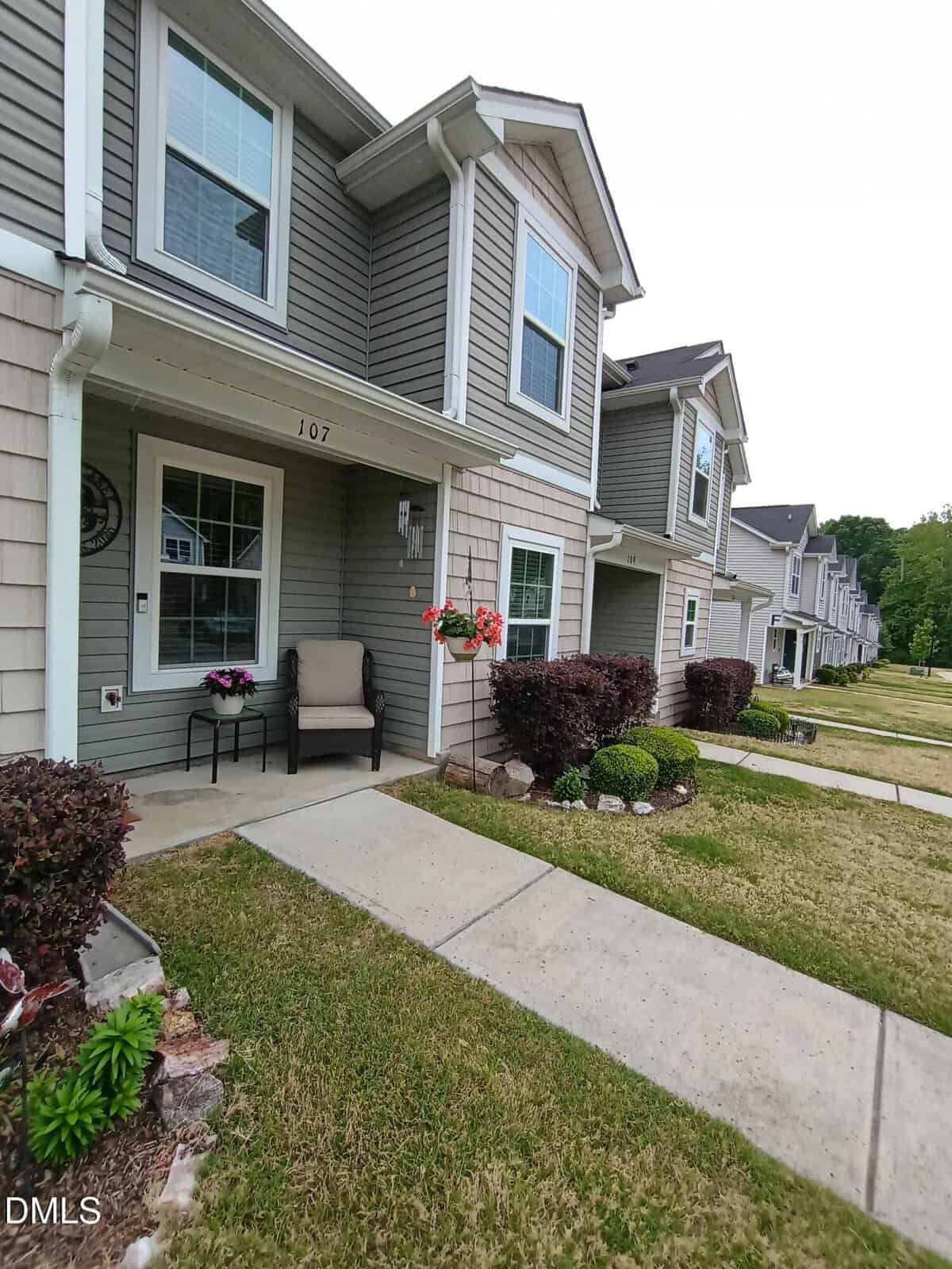 Two-story townhouse with gray siding, white trim, and a front porch with a chair and flowers. Well-kept bushes and a sidewalk line the front, with similar homes visible down the row.