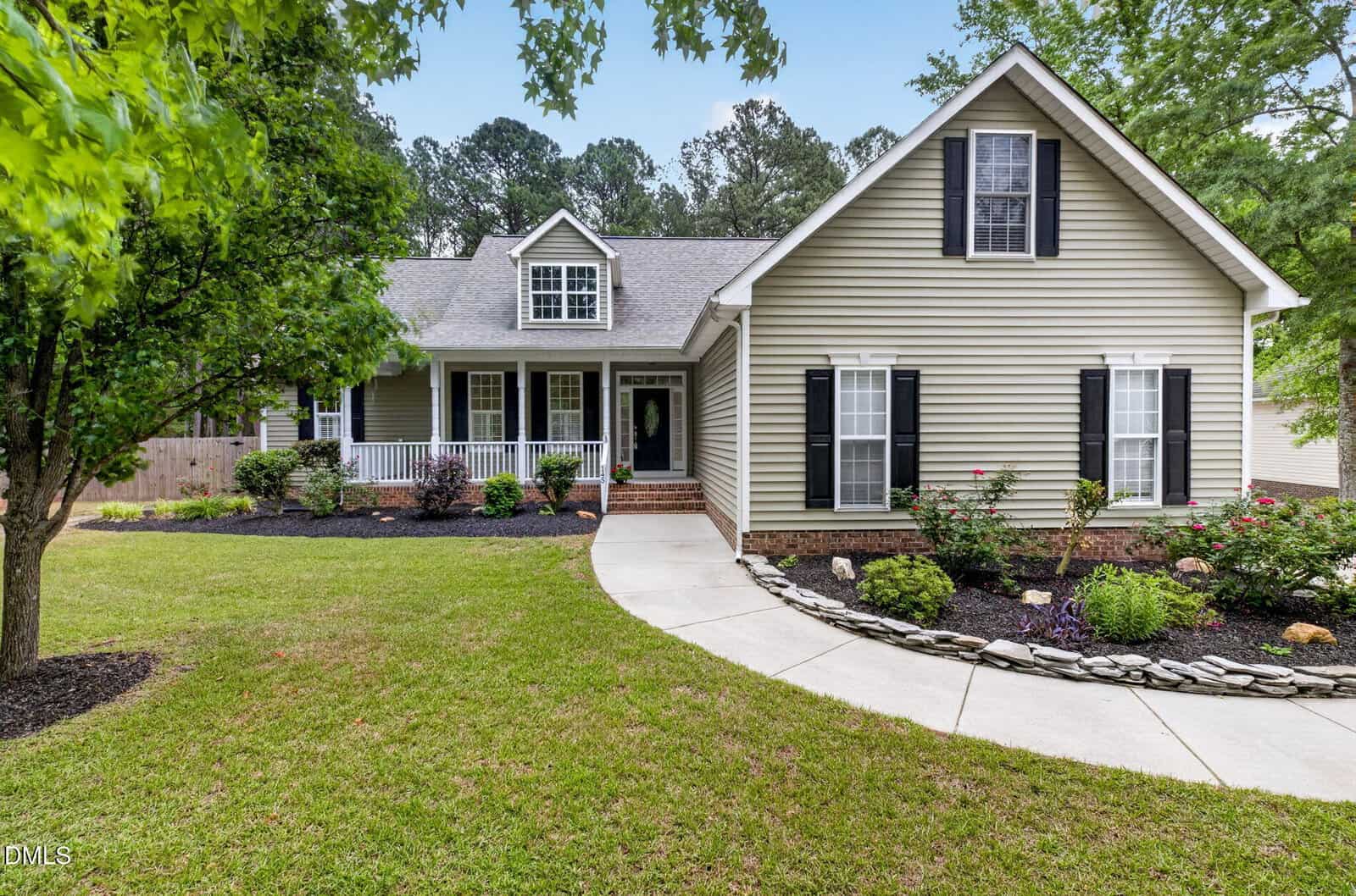 A light green house with black shutters, a front porch with white railings, and a dormer window. The yard has green grass, shrubs, flowers, and a curved walkway leading to the front door.