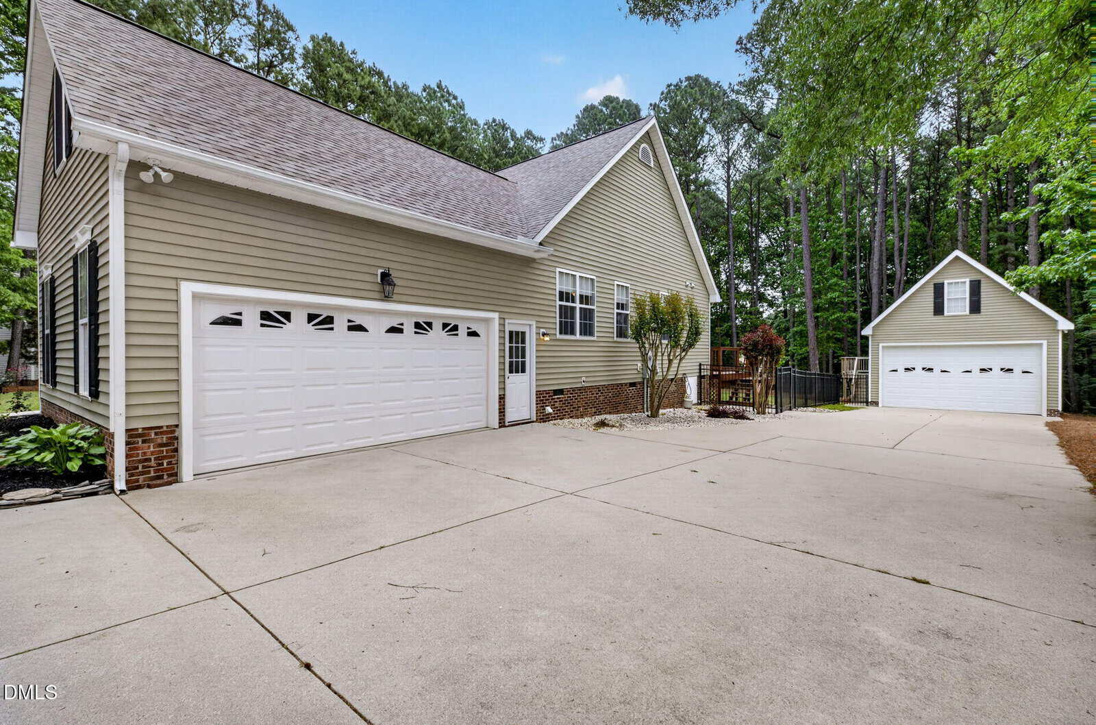A large beige house with two white garage doors and a spacious concrete driveway, next to a separate detached garage, surrounded by trees and greenery.