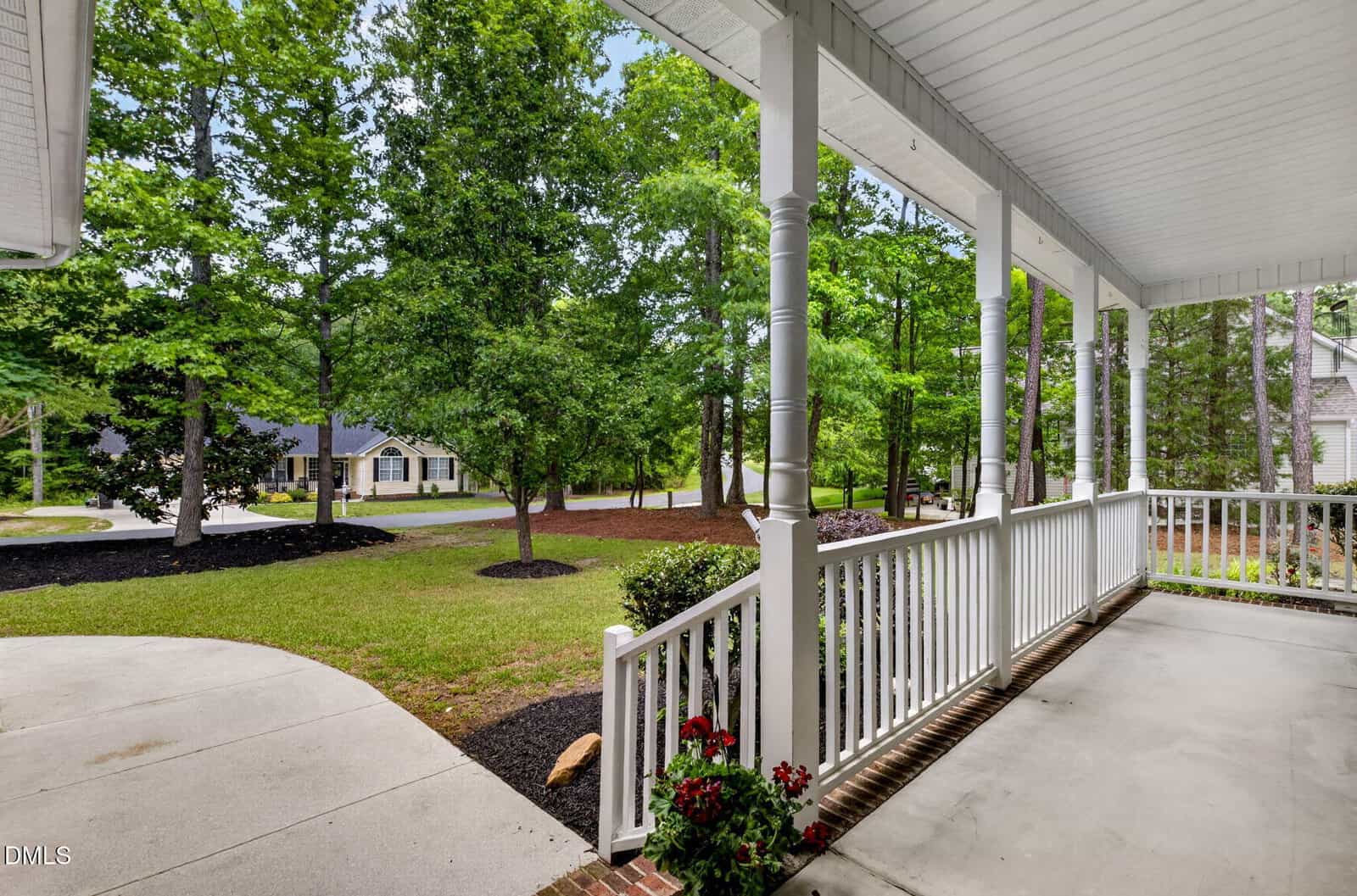 Covered front porch with white railing and columns, overlooking a landscaped yard with green grass, mulch, and tall trees. A few red flowers are in a pot by the porch. Houses and a quiet street are visible in the background.