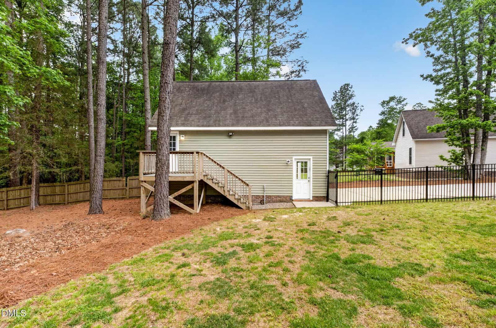 A small, light-colored house with a sloped roof, wooden stairs, and a deck sits next to a fenced yard and tall trees, surrounded by green grass under a clear blue sky.