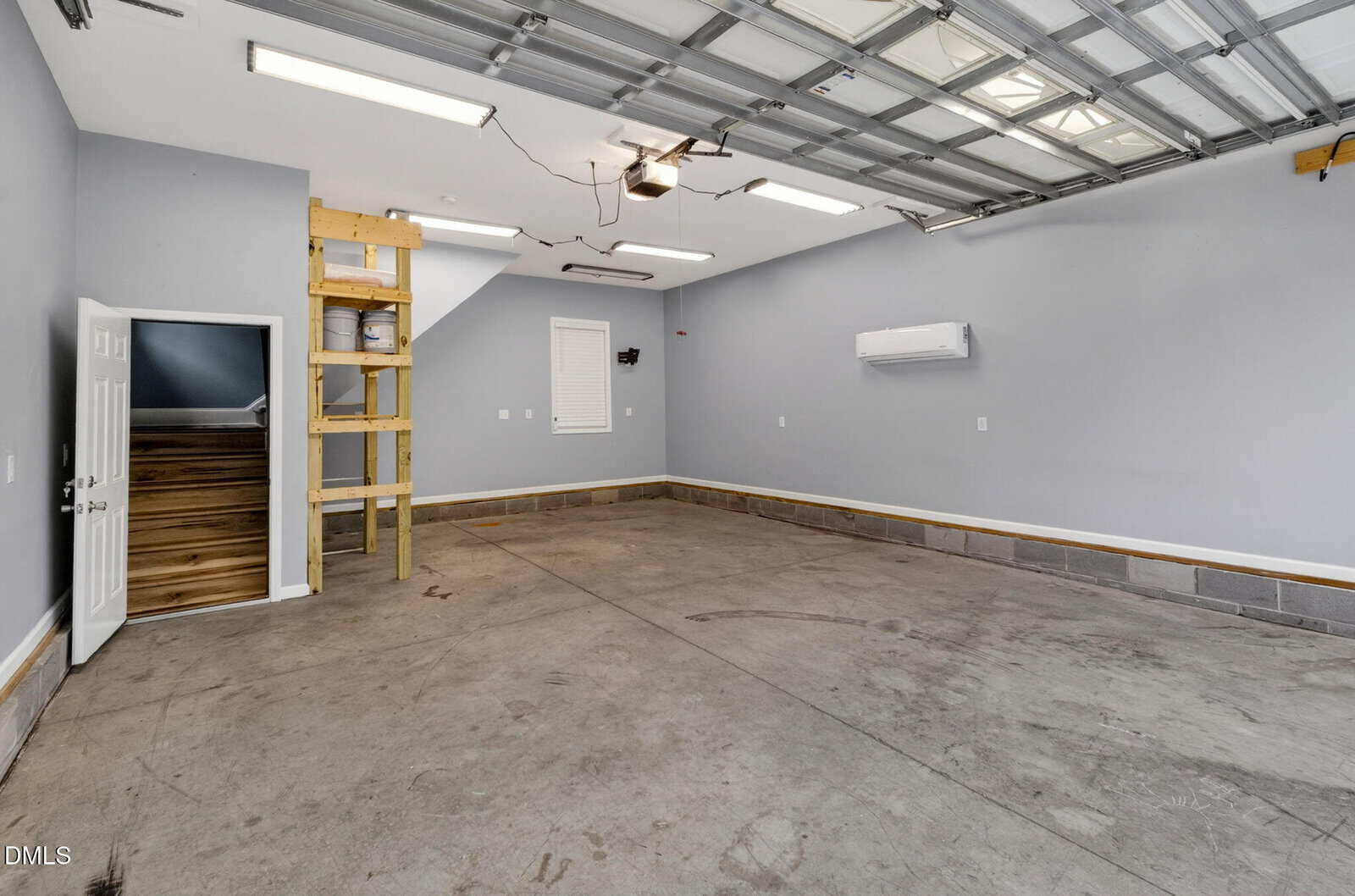 A spacious, clean two-car garage with gray walls, concrete floor, a mini-split AC unit, open ceiling with lights, a ladder to a loft, and a door leading to a staircase.