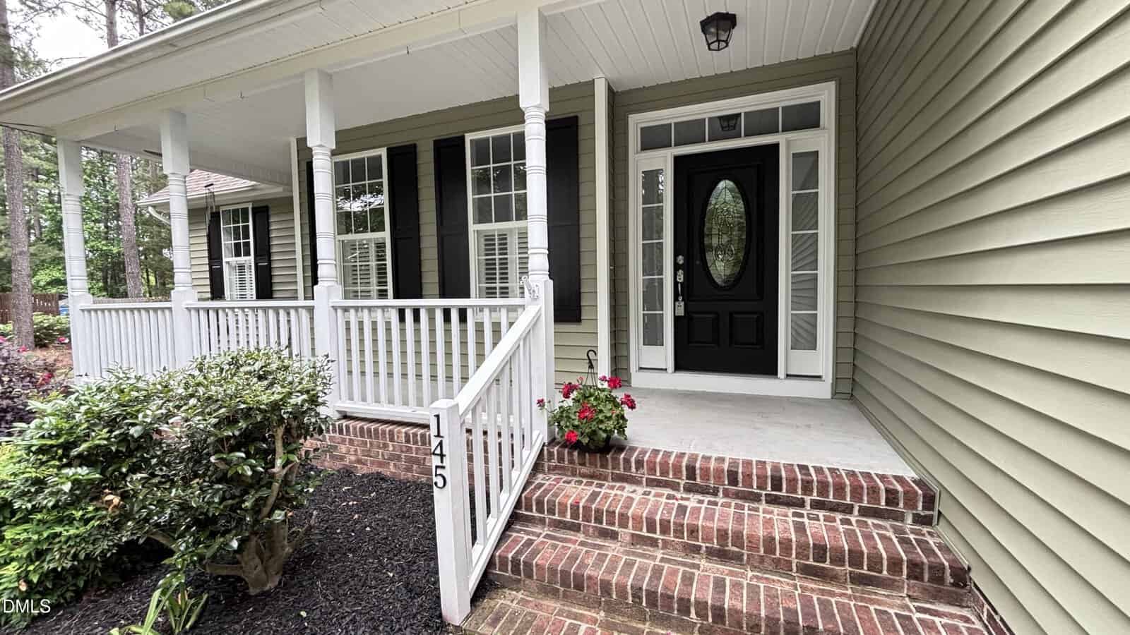 Front view of a house with green siding, black shutters, white columns, a covered porch, brick steps, a black front door with glass panels, and potted flowers beside the entrance.
