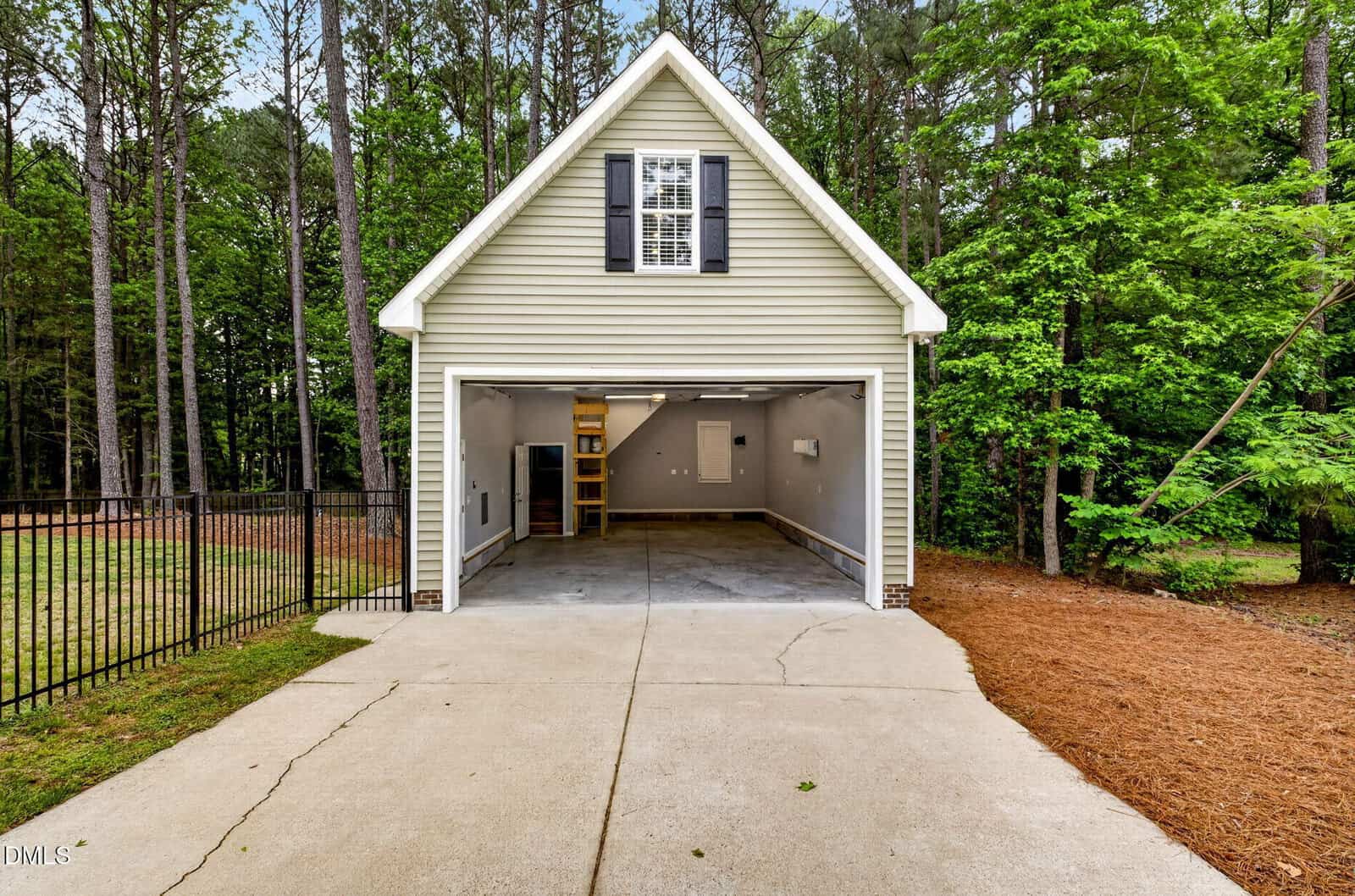 A two-car garage with open doors, attached to a house surrounded by tall trees and greenery. The driveway is concrete, and the garage interior appears clean and empty with shelves and stairs inside.