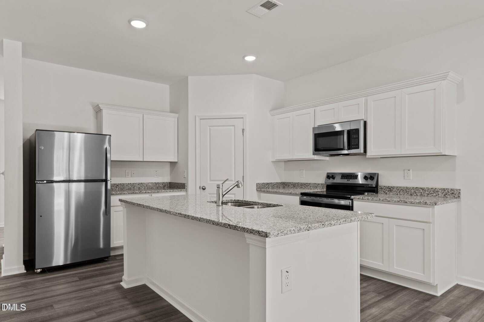 Modern kitchen with white cabinets, granite countertops, stainless steel refrigerator, oven, and microwave, featuring a large island with a sink and wood-look flooring. The space is clean and well-lit.