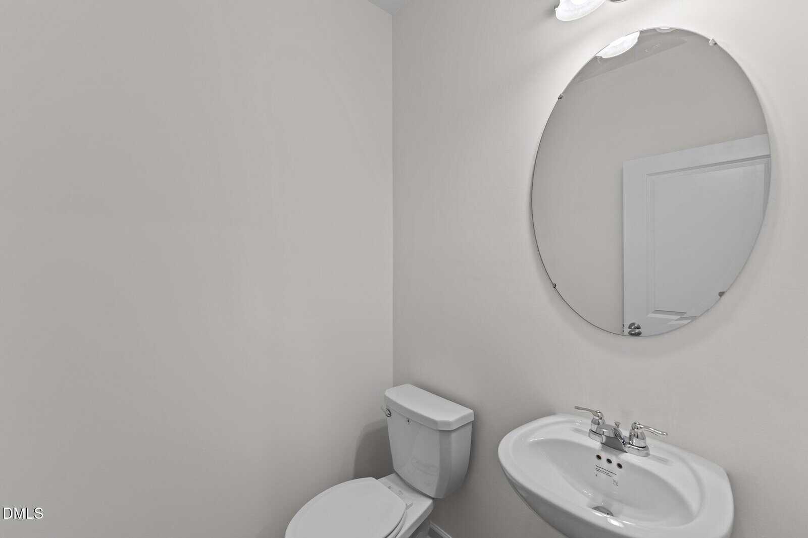 Minimalist bathroom with white walls, a toilet, a small sink with chrome fixtures, and a round mirror above the sink. The door is visible in the mirror’s reflection.