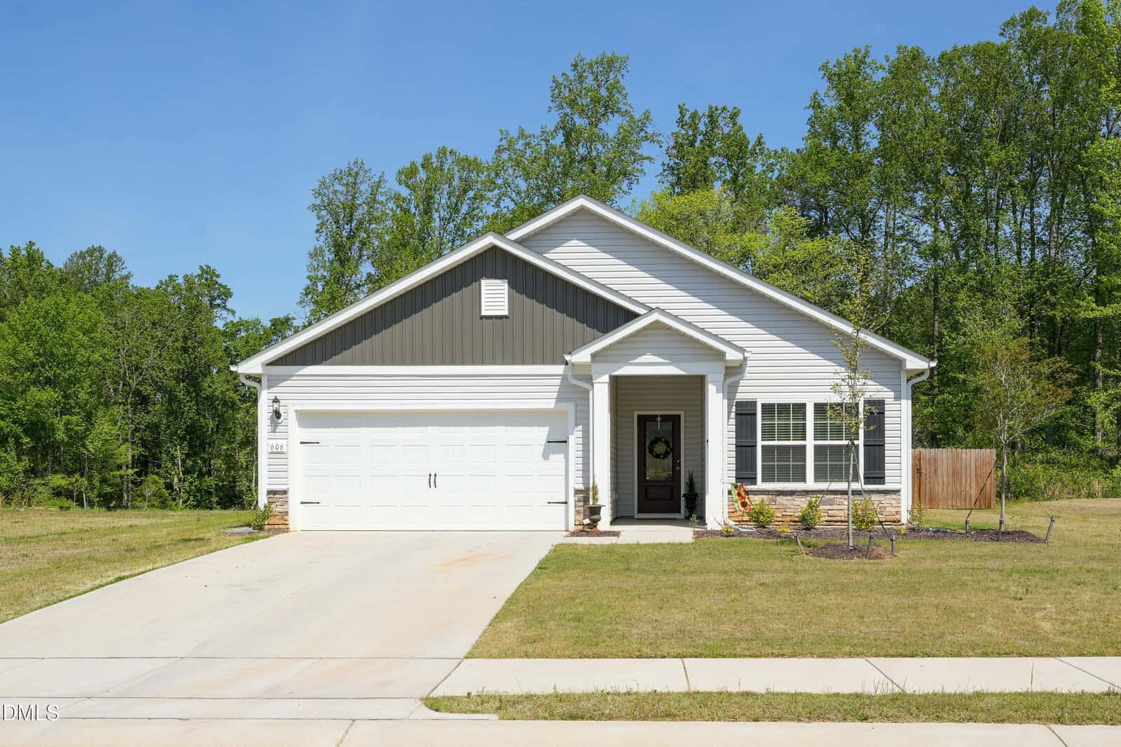 Single-story suburban house with gray siding and white trim, attached two-car garage, small covered porch, and simple landscaping; surrounded by grass and trees under a clear blue sky.