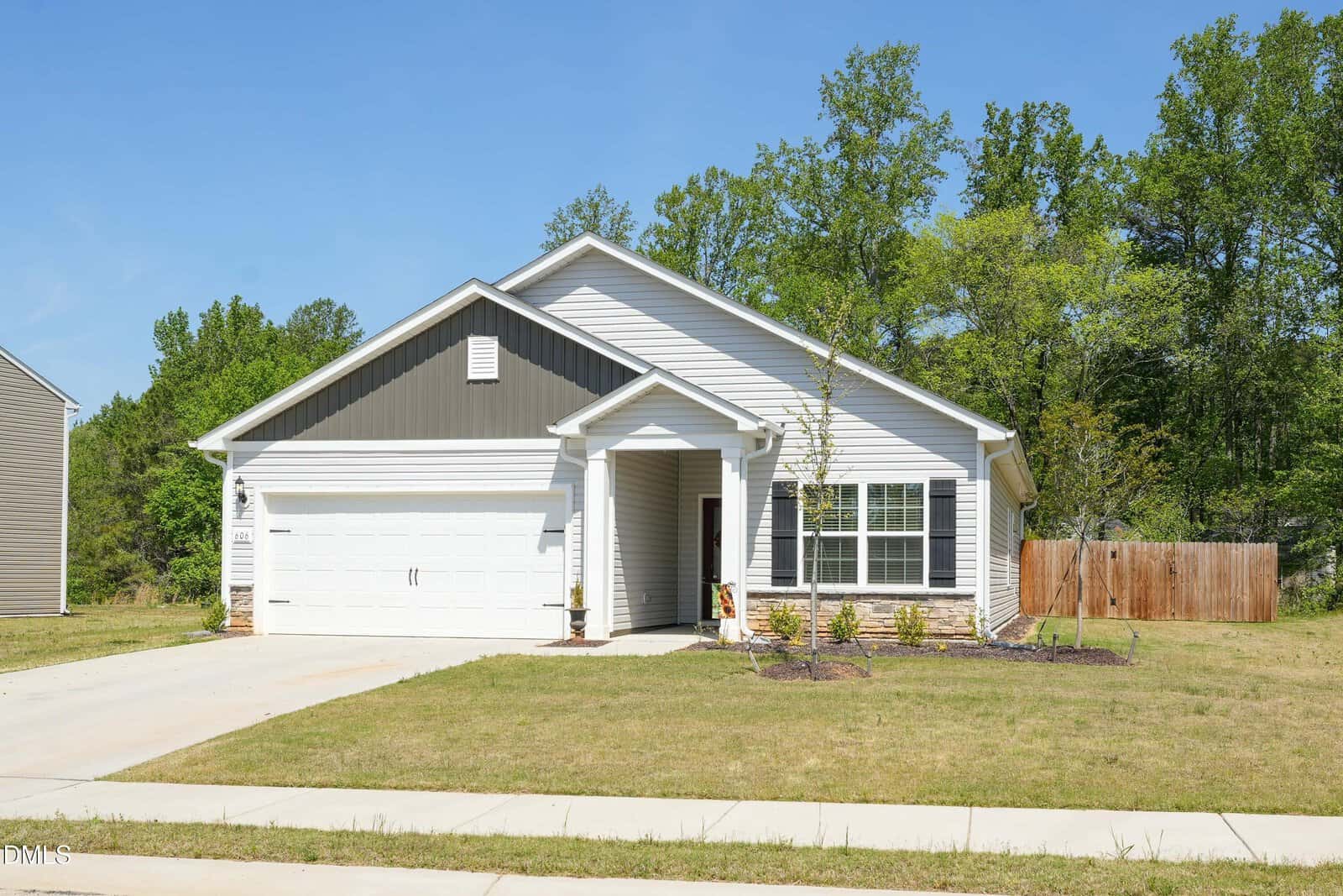 Single-story suburban house with white siding, gray gable, and attached two-car garage, set on a well-kept lawn with small plants by the entrance and trees in the background under a clear blue sky.