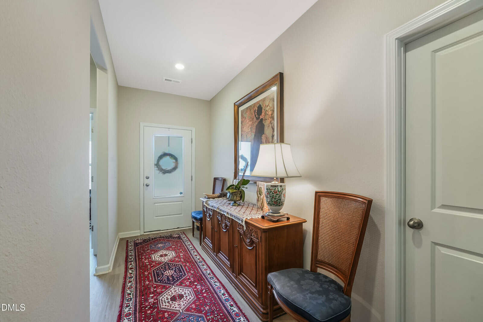A hallway with a red patterned rug, a wooden sideboard topped with a lamp and decor, a chair, light-colored walls, and a glass door with a wreath at the end. Artwork hangs above the sideboard.