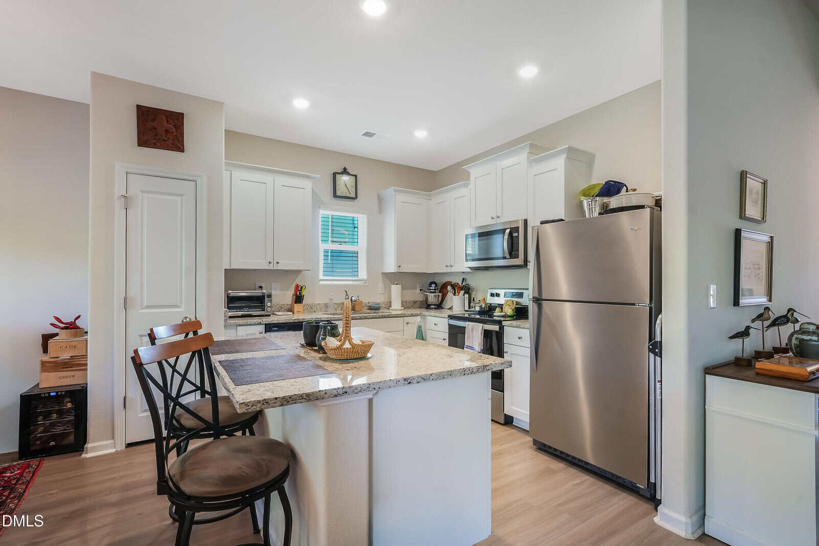 A modern kitchen with white cabinets, stainless steel appliances, a center island with two chairs, and various kitchen items on the counters. Light wood flooring and neutral-colored walls create a bright, inviting space.