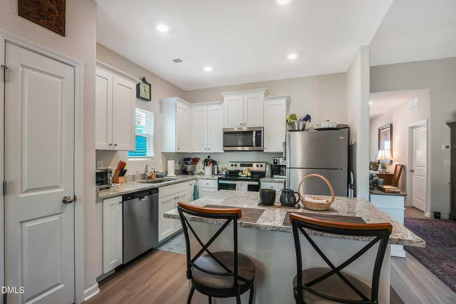 Modern kitchen with white cabinets, stainless steel appliances, and a granite island with two chairs. The counter has a basket and teapot, and the room is well-lit with recessed lighting. A hallway leads to another room.