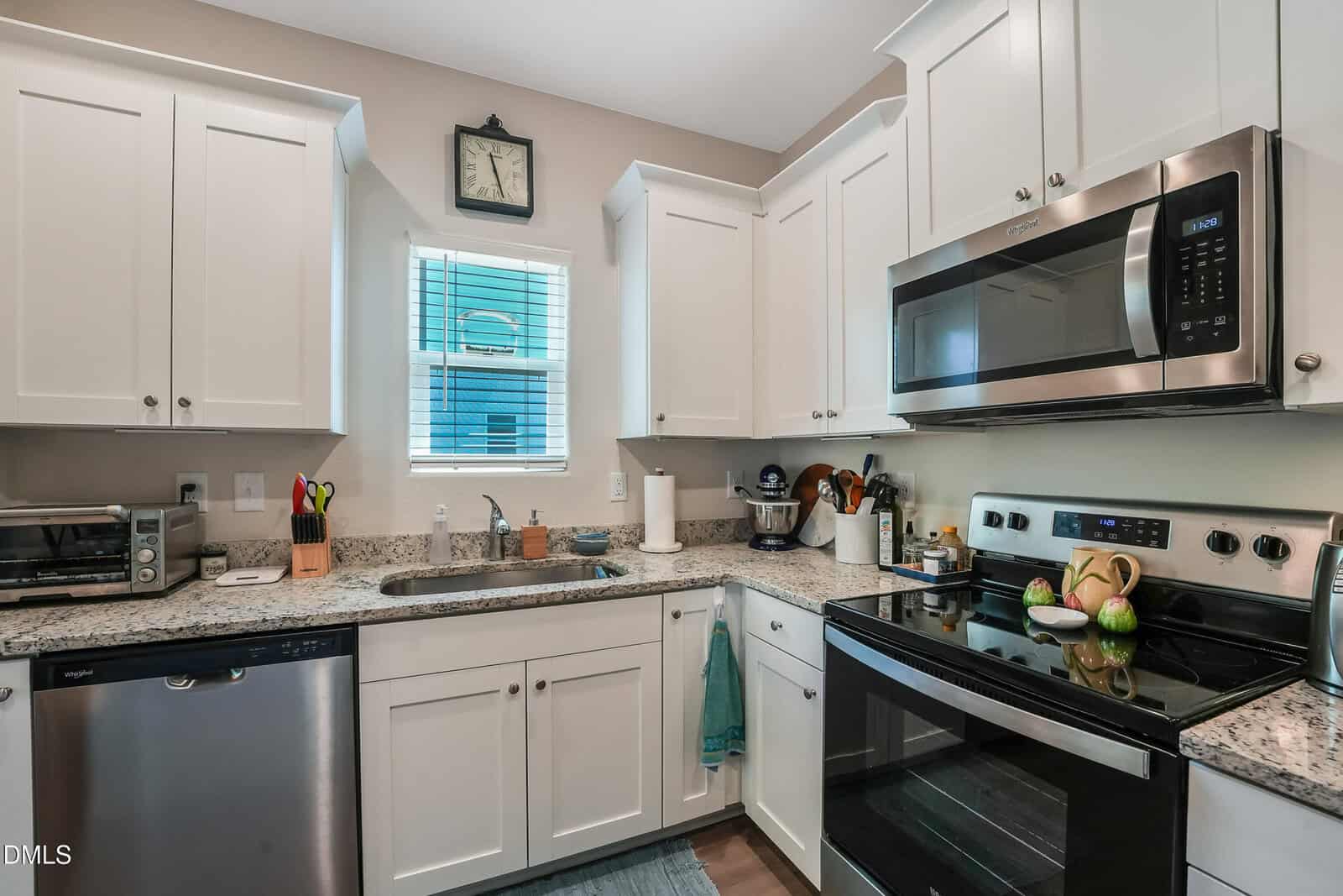 Modern kitchen with white cabinets, granite countertops, stainless steel appliances, a window above the sink, and various kitchen tools on the countertops. A clock hangs above the window.