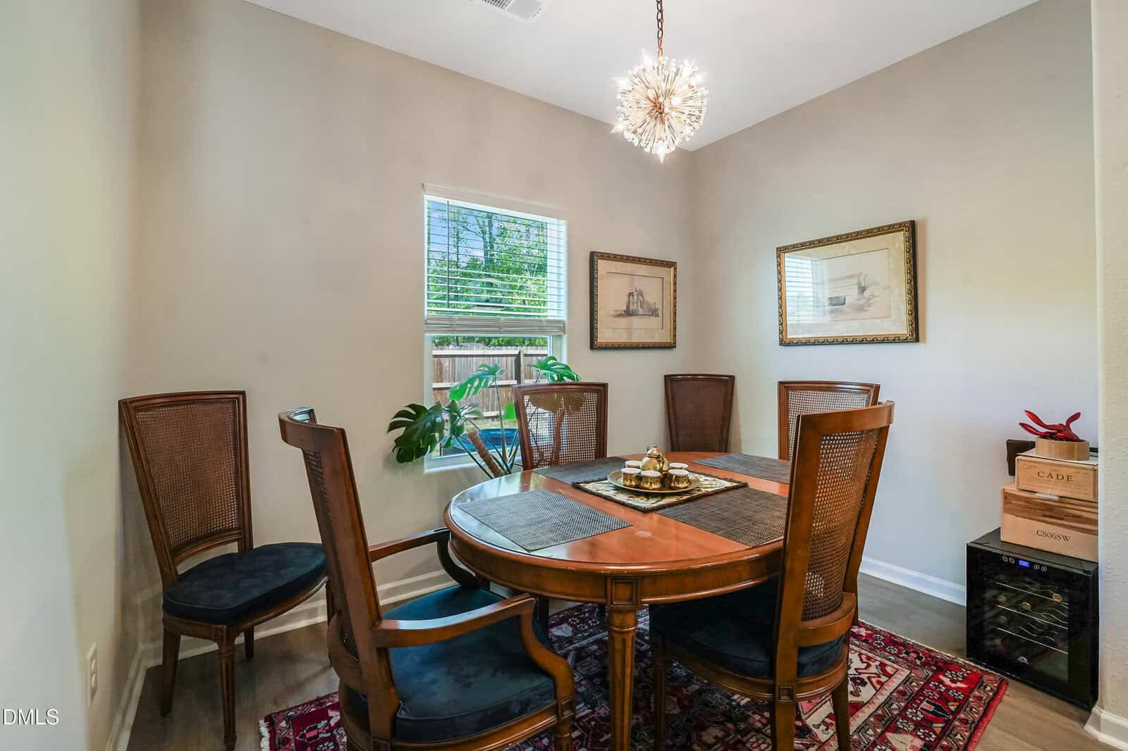 A dining room with a round wooden table, six chairs, a plant by the window, framed art on beige walls, a modern chandelier, and a patterned rug on the floor.