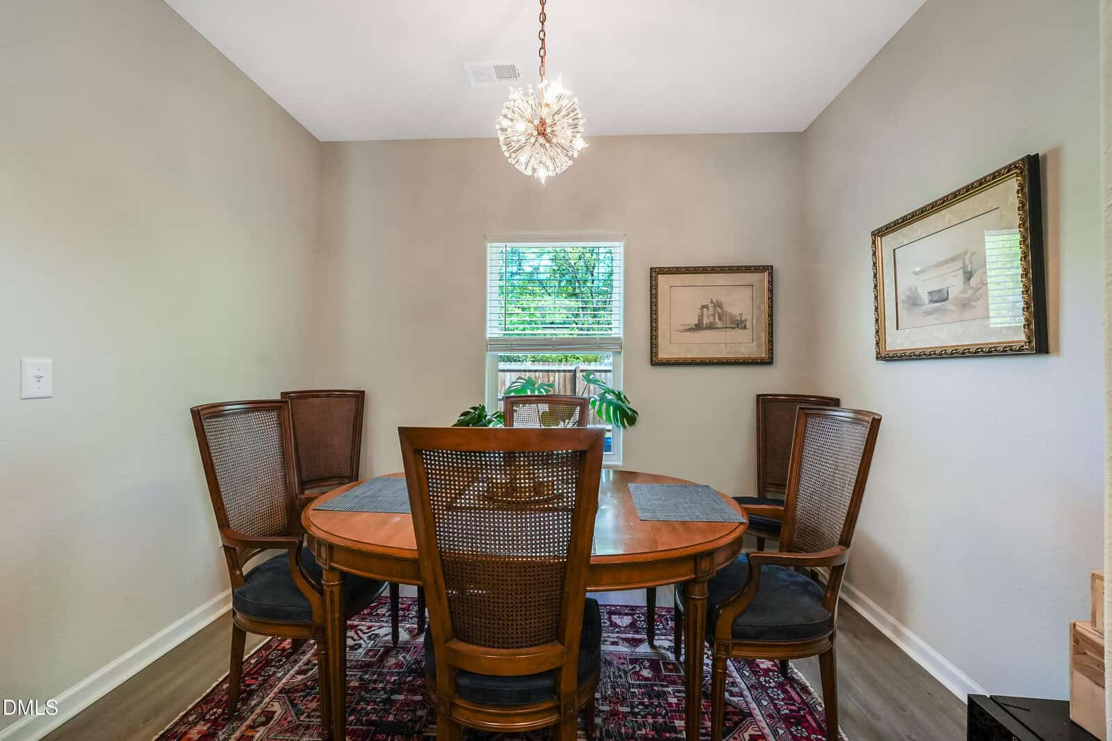 A dining room with a wooden table, six chairs, a patterned rug, two framed artworks on beige walls, a window with blinds, a green plant, and a chandelier overhead.