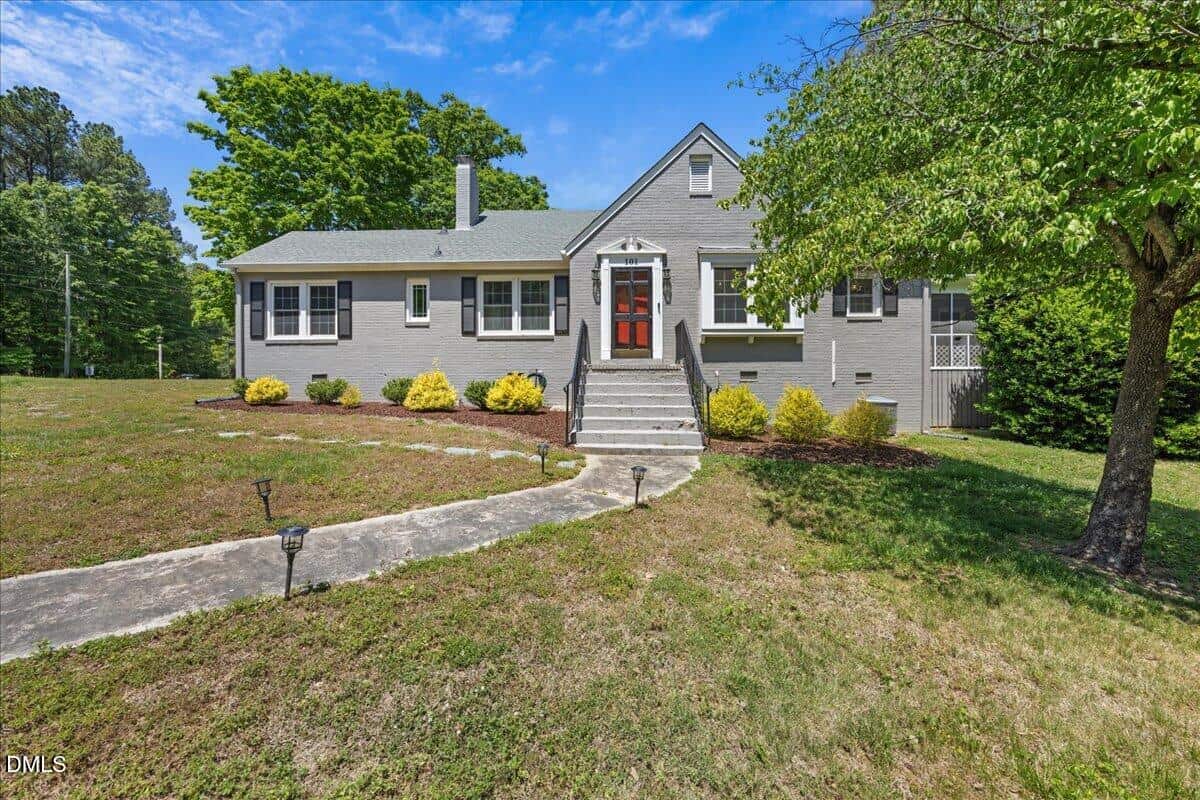 A gray single-story house with white trim and a red front door. The house has a concrete path, steps leading to the entrance, small bushes along the front, and a large grassy yard with a tree on the right.